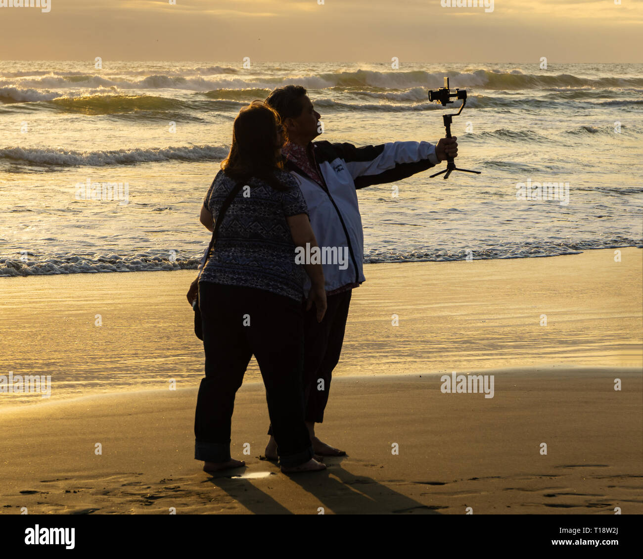 Two adult people vlogging on the beach at sunset. Man holding his camera to vlog. Cannon Beach, OR, USA. Stock Photo