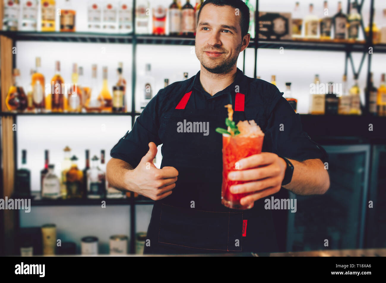 Nice and young barman is holding a glass of cold cocktail in the left hand and showing the big thumb up with the right one. He recommends this cocktai Stock Photo
