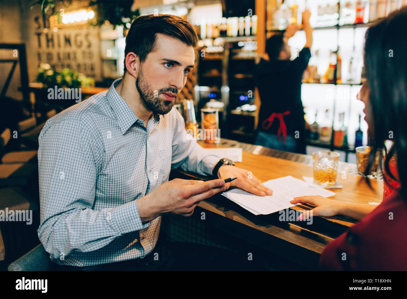 A business meeting of two people in a bar. Man is explaining something to woman. She is listening to him very accurate and careful. Barman is standing Stock Photo