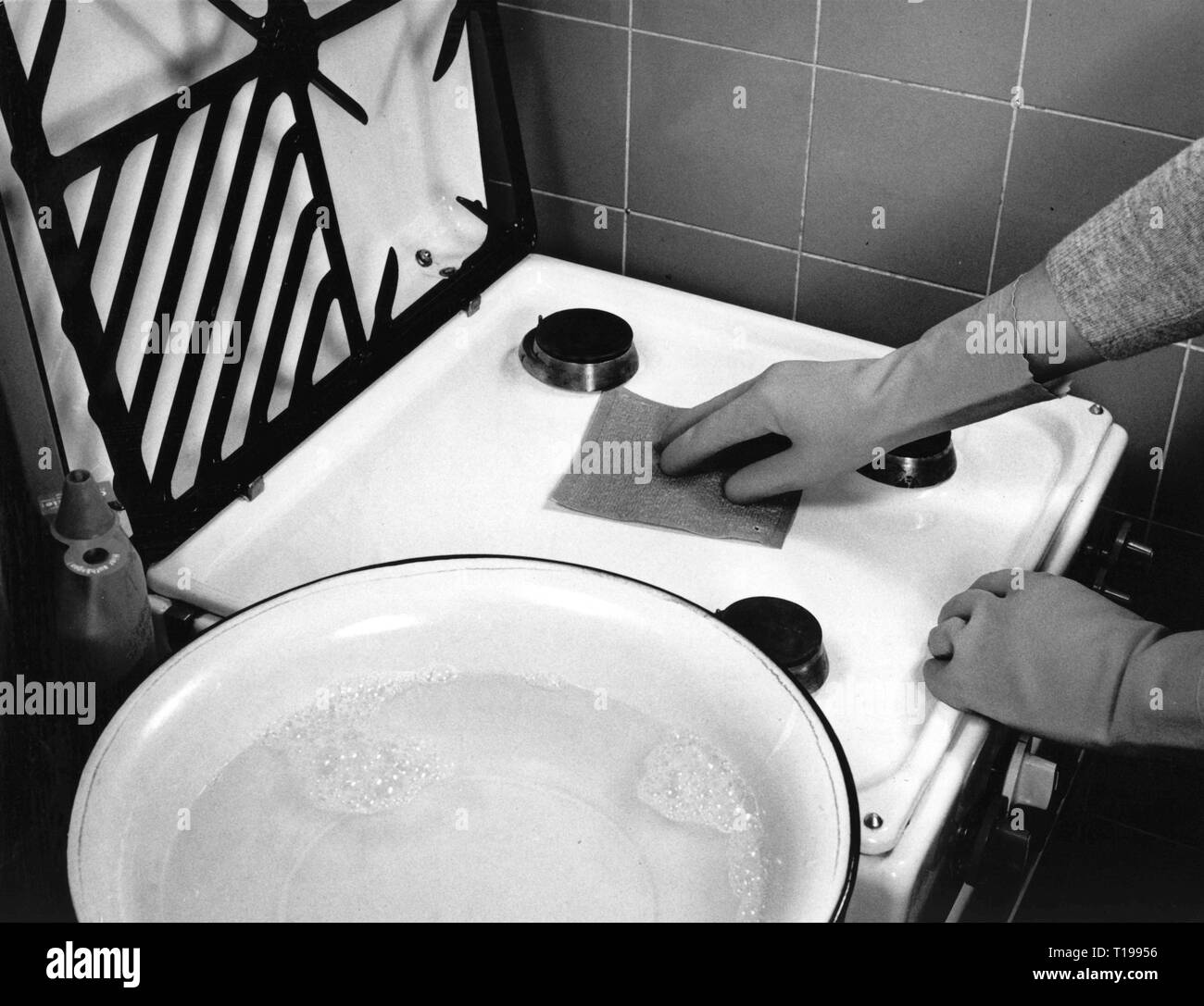 household, kitchen and kitchenware, cleaning a gas stove, 1960s, range, stoves, hob, stove, stove top, stovetop, cook, cooking, cleanliness, cleanse, cleansing, cloth, cloths, rubber glove, rubber gloves, glove, gloves, household chores, do the chores, housekeep, domestic work, housework, cooker, cookers, water bowl, bowl, bowls, water, enamel, household, households, kitchen, kitchens, cleaning, clean, gas stove, gas stoves, historic, historical, people, 20th century, Additional-Rights-Clearance-Info-Not-Available Stock Photo