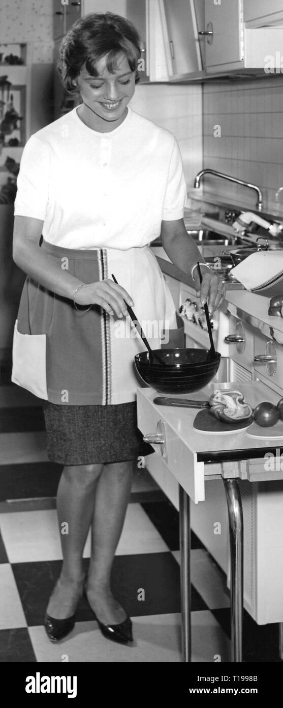 household, cooking and baking, young housewife preparing a salad, 1960s, household chores, do the chores, housekeep, domestic work, housework, half-length, half length, standing, apron, aprons, smiles, smiling, smile, holding, hold, salad servers, salad bowl, salad bowls, bowl, bowls, mix, mixing, table, tables, kitchen, kitchens, kitchen unit, cupboards, wall cupboard, stove, stoves, cooker, cookers, kitchen sink, kitchen sinks, household, households, cook, cooking, bake, baking, housewife, housewives, homemaker, preparation, preparations, salad, Additional-Rights-Clearance-Info-Not-Available Stock Photo