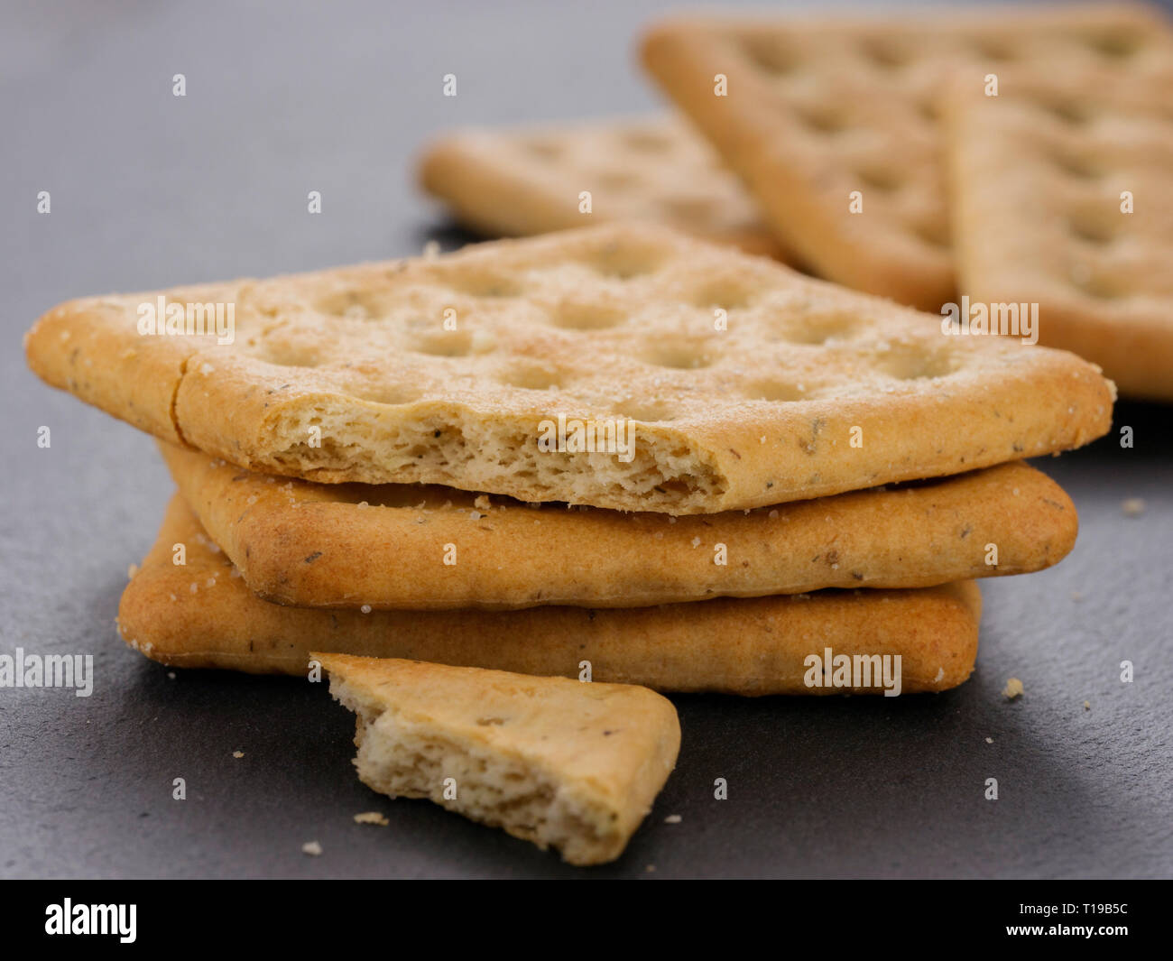 stack of square crackers with pieces and crumbs on slate gray background. Dry salt cracker cookies with fiber and dry spices Stock Photo