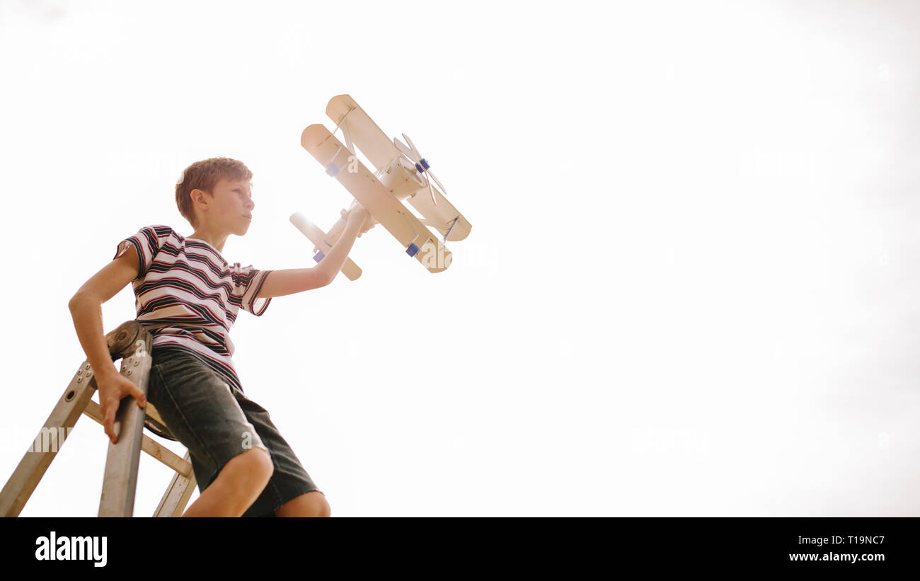 Boy sitting on top of a ladder with toy airplane against bright sunlight. Boy imagining to be pilot, flying a top aeroplane. Stock Photo