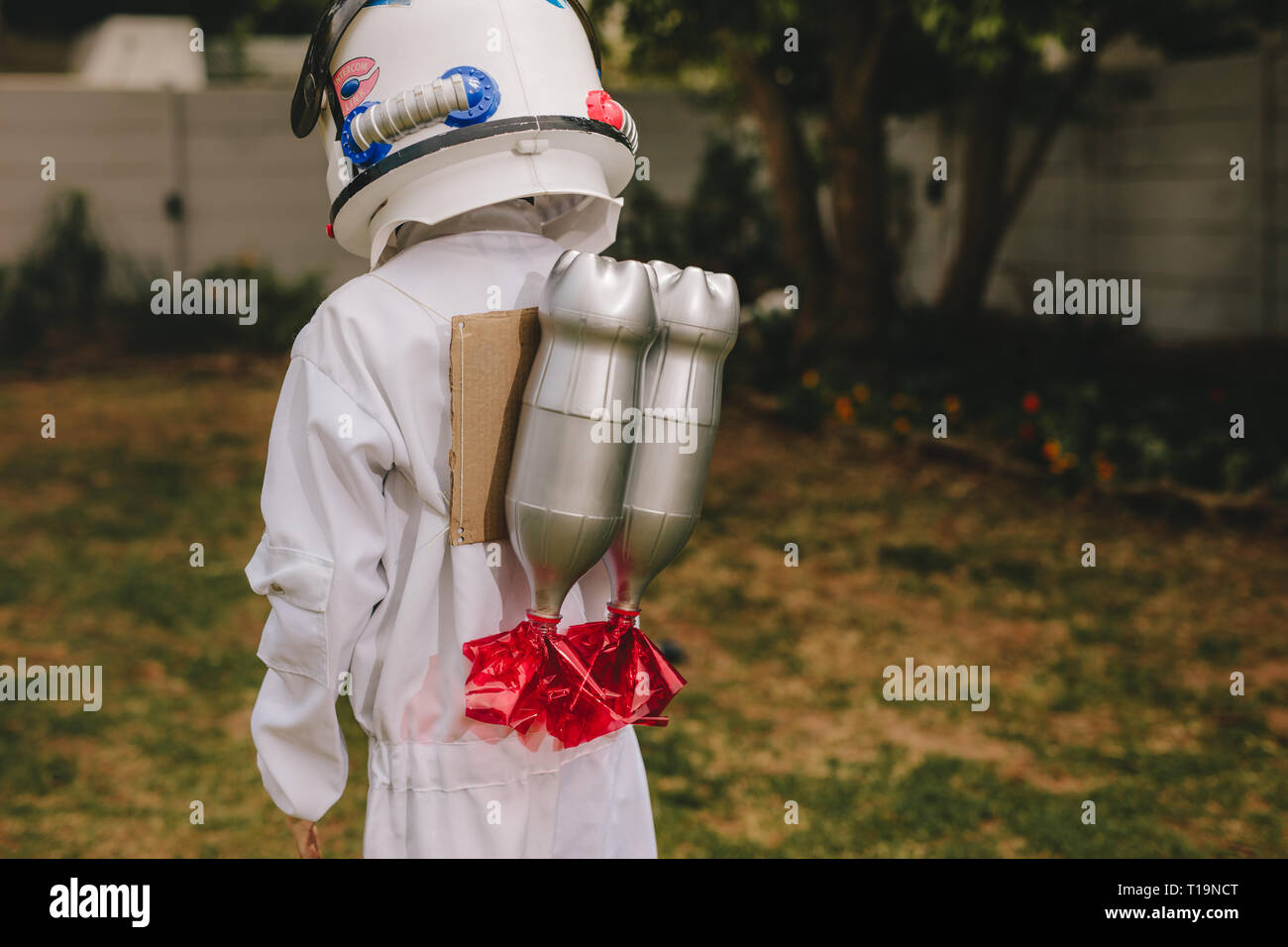 Rear view of boy in space helmet and suit carrying a toy jetpack on his back. Boy pretending to be an astronaut playing outdoors. Stock Photo