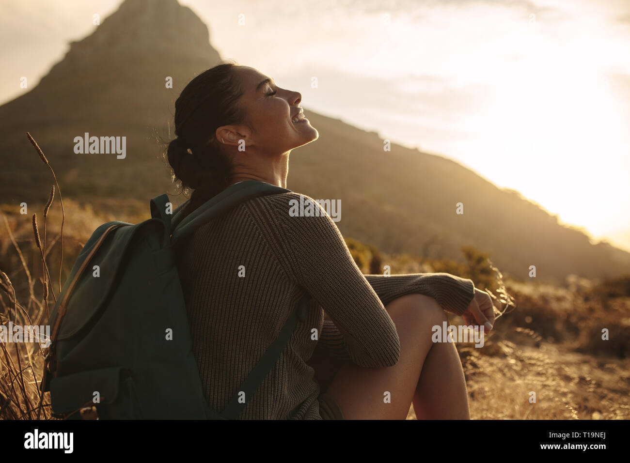 Woman sitting on ground smiling with her eyes closed. Caucasian female tourist taking a break after hiking on country trail. Stock Photo