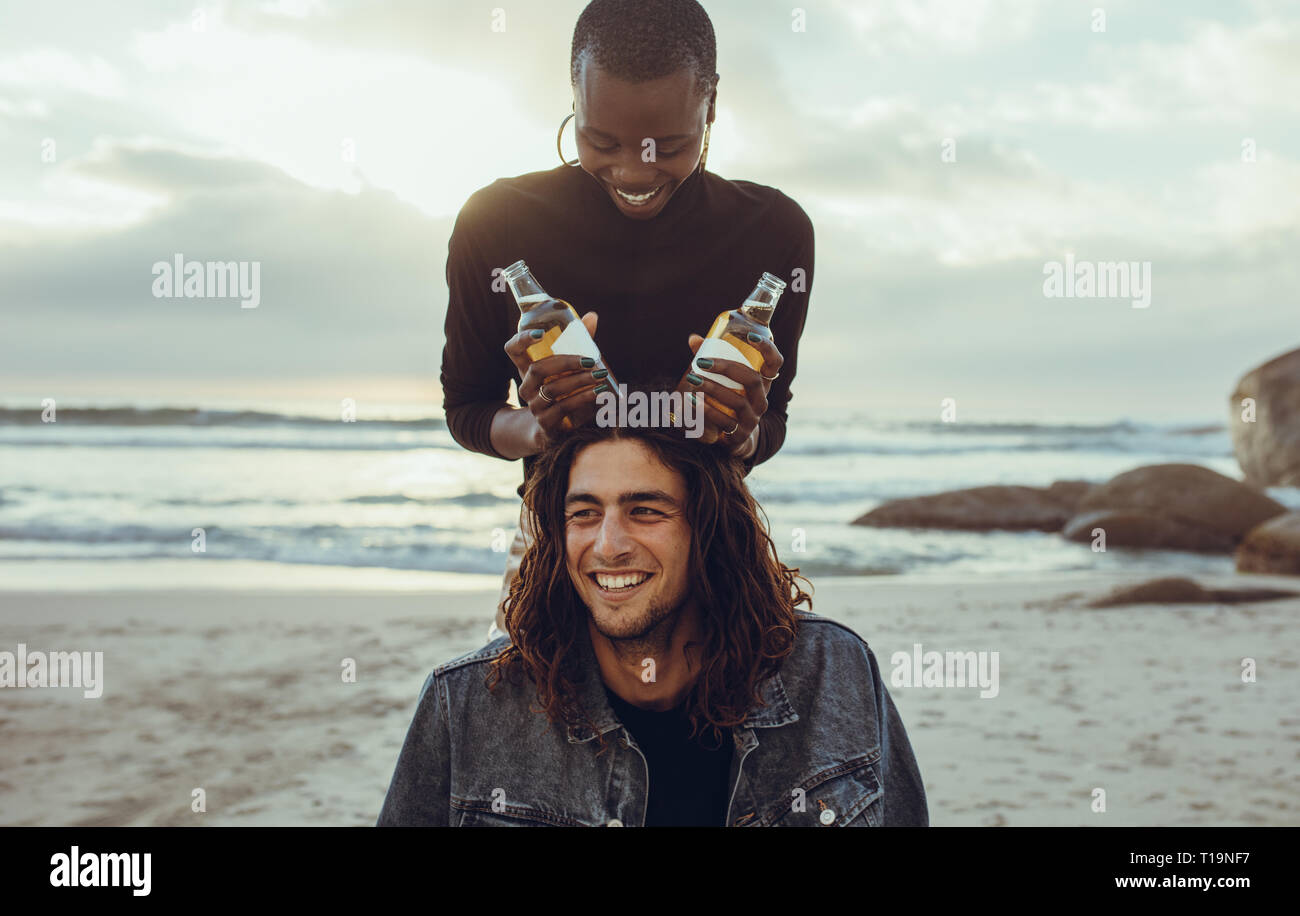 Woman holding beer bottles on the head of men as horns. Couple of friends enjoying themselves at the beach. Stock Photo