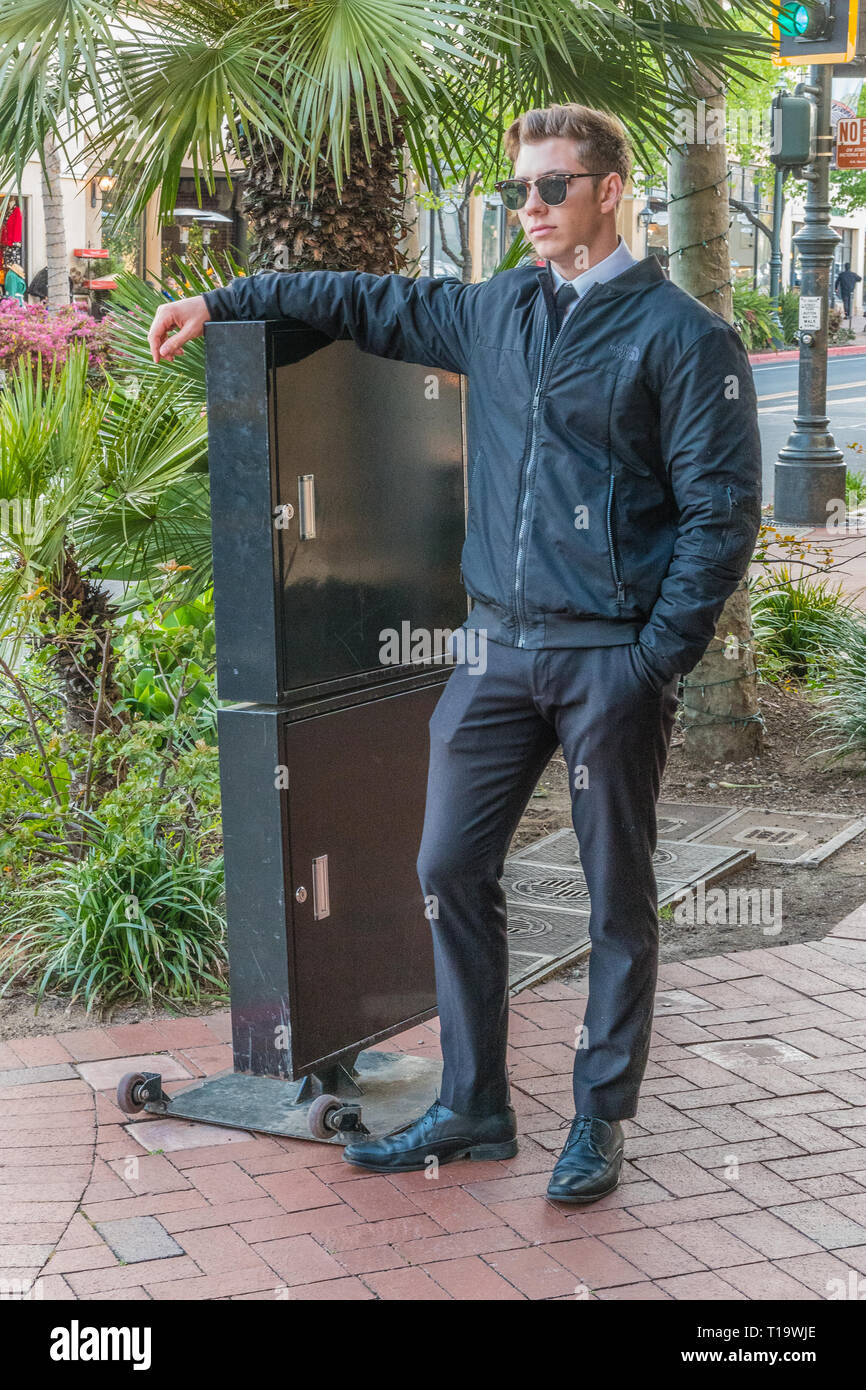 A young adult valet, dressed in all black, stands with one arm on his