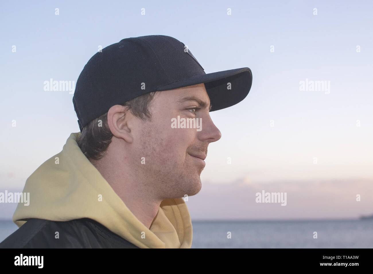 Profile shot of handsome smiling natural and casual looking male in late twenties with hood jacket and cap looks straight forward, ocean horizon behin Stock Photo