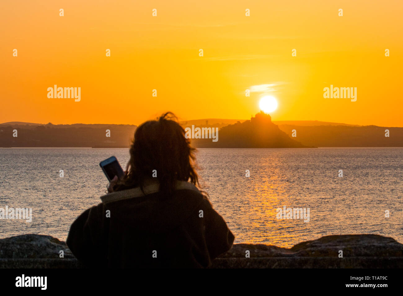 Penzance, Cornwall, UK. 25th Mar, 2019. UK Weather. The sunrises in clear skies, above St Michaels Mount in Cornwall. An early morning walker pauses to take a picture on her mobile phone. Credit: Simon Maycock/Alamy Live News Stock Photo