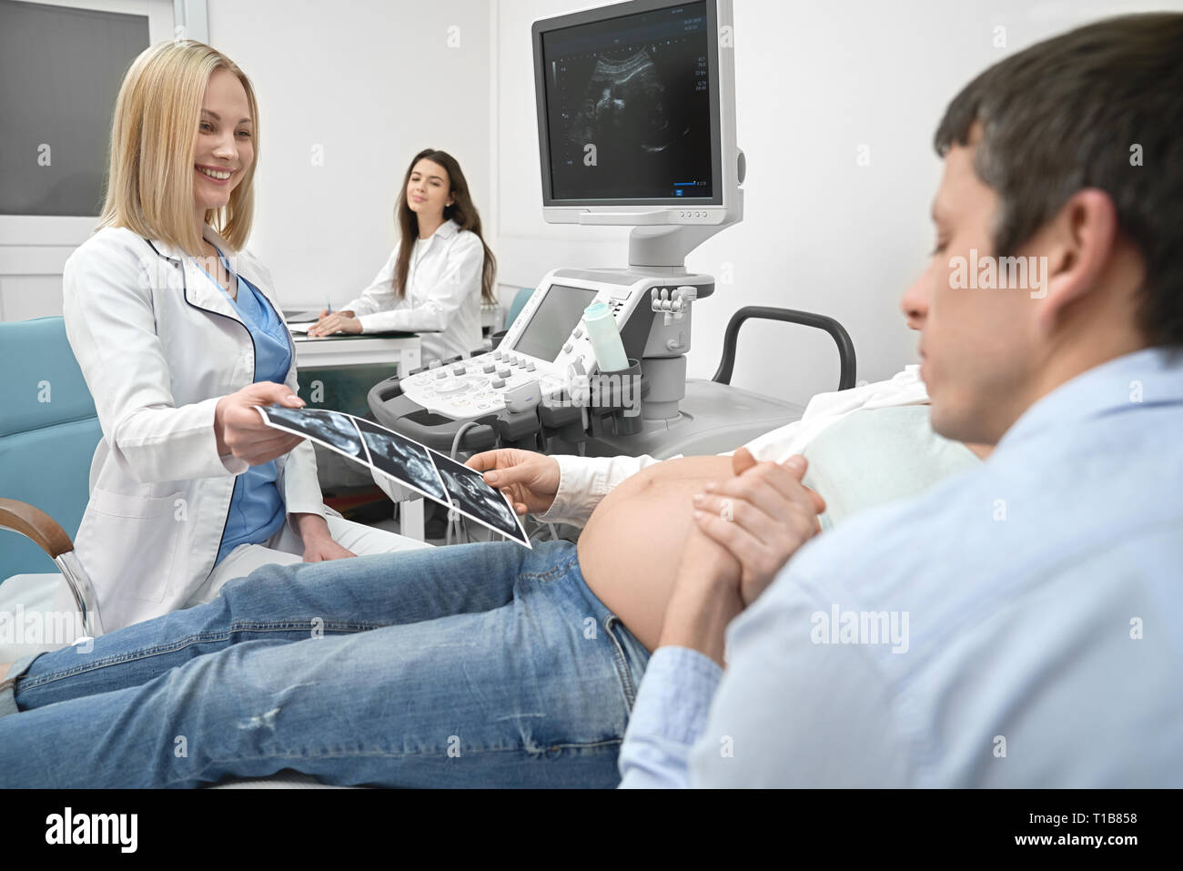 Husband and wife on ultrasound examination in medical clinic. Man holding hand of pregnant woman, future parents looking at x ray. Female doctor, beautiful blonde woman showing sonogram. Stock Photo