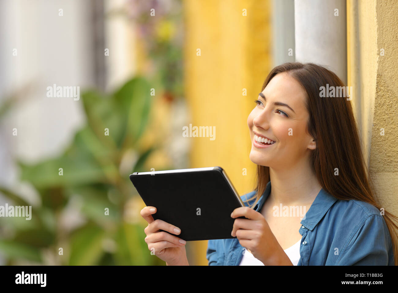 Happy woman holding tablet thinking looking at side in a colorful street Stock Photo