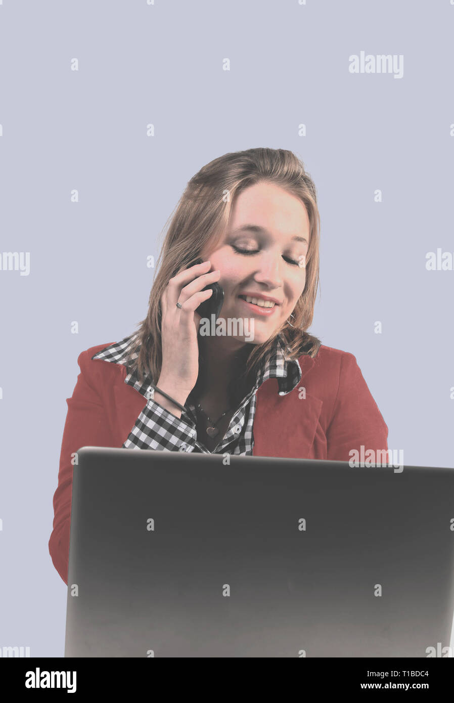 Young woman with red jacket answering phone while looking at laptop Stock Photo
