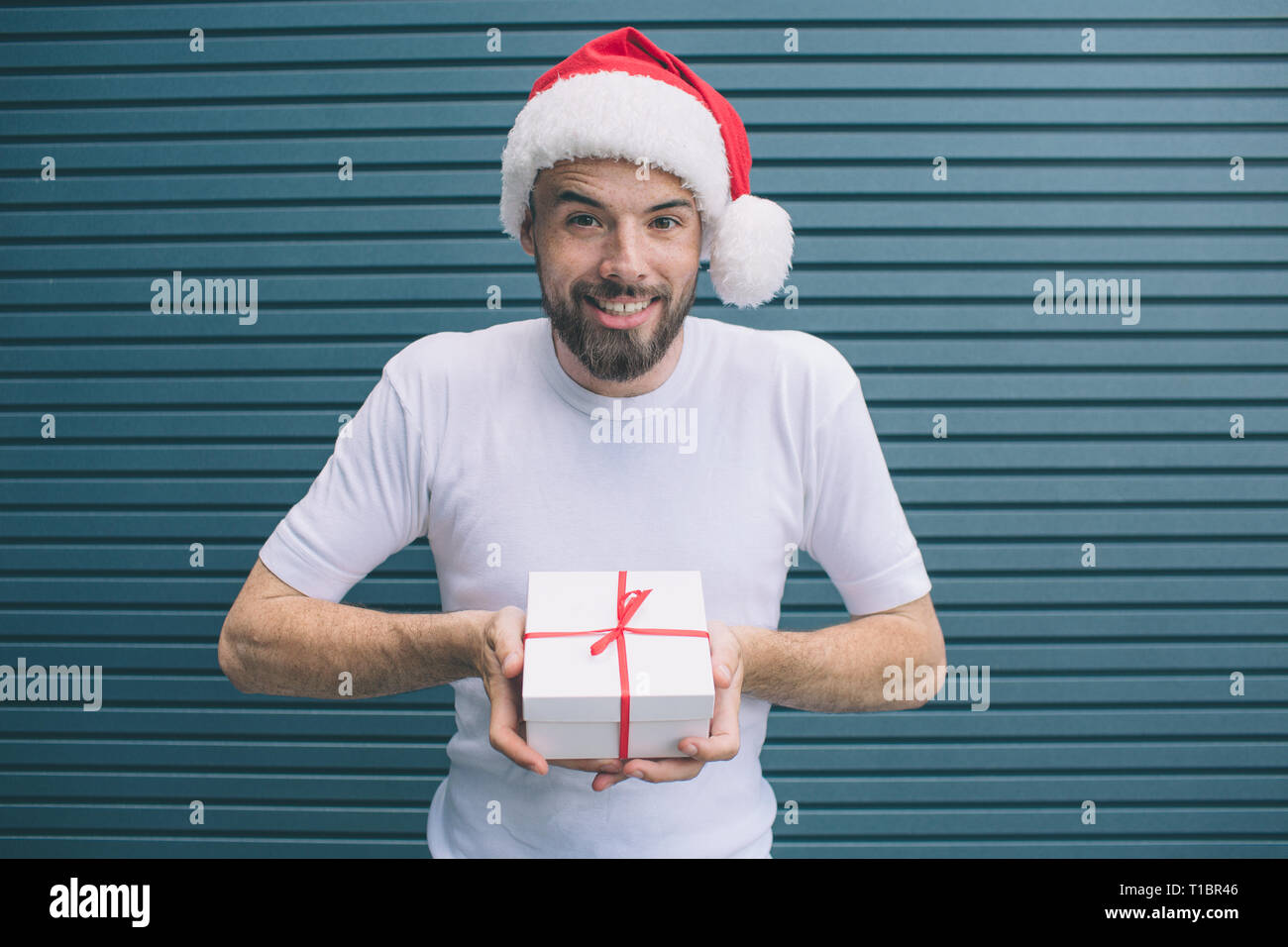 Delightful man in Christmas hat is standing and holding present in white cover. He is looking forward on camera. Guy is smiling. He is very positive Stock Photo