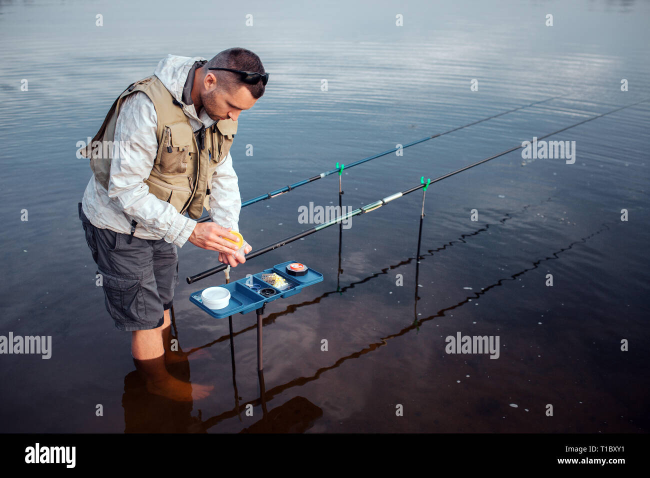 Young fisherman is standing in water barefoot. He is leaning to opened plastic box with artificial baits. There are two fly rods lying in hooks. Stock Photo