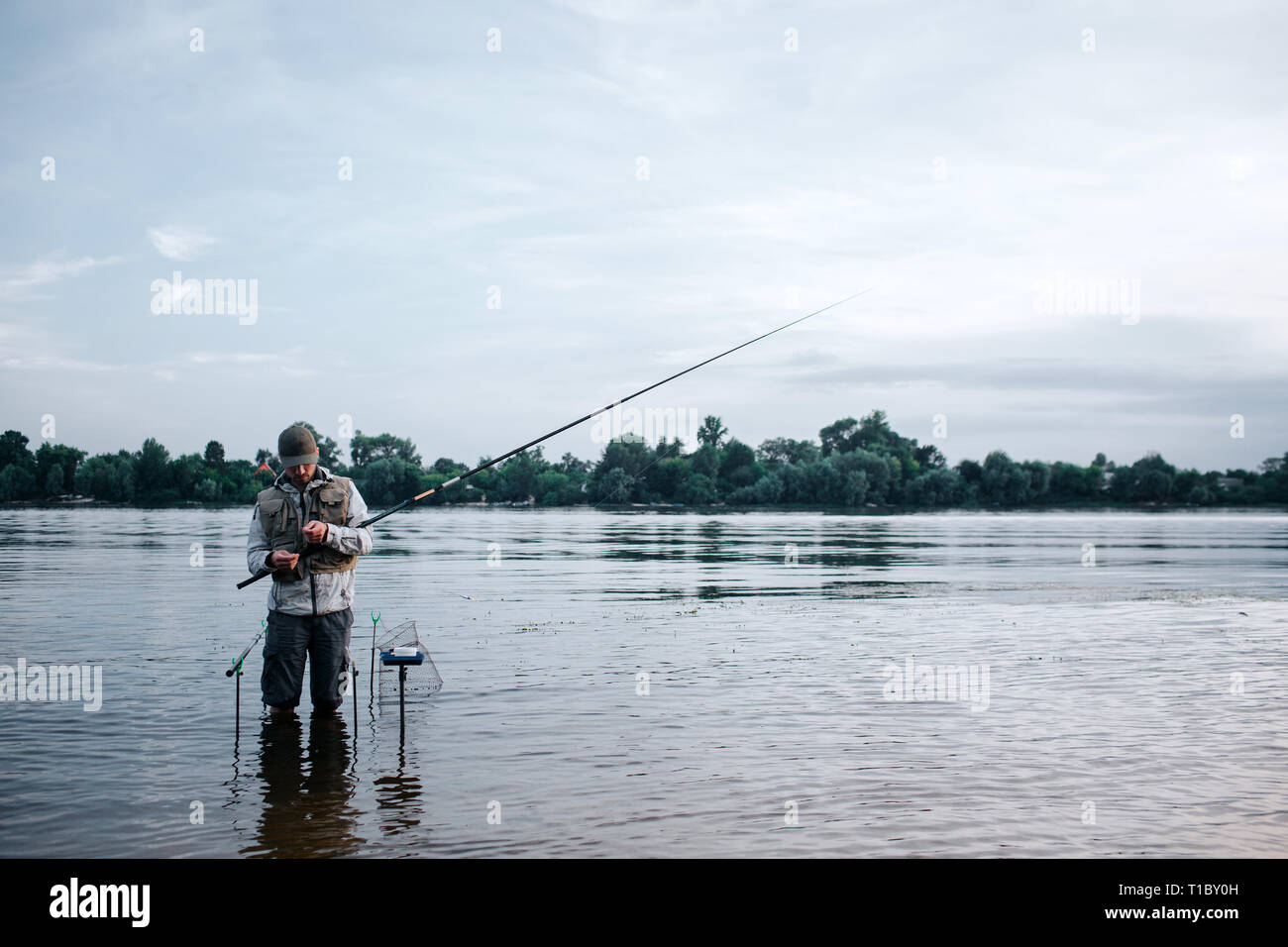 Young fisherman is standing barefeet in water and holding fly rod. He looks  at it. Guy is working with spoon and bait. He stands near hooks that put  Stock Photo - Alamy