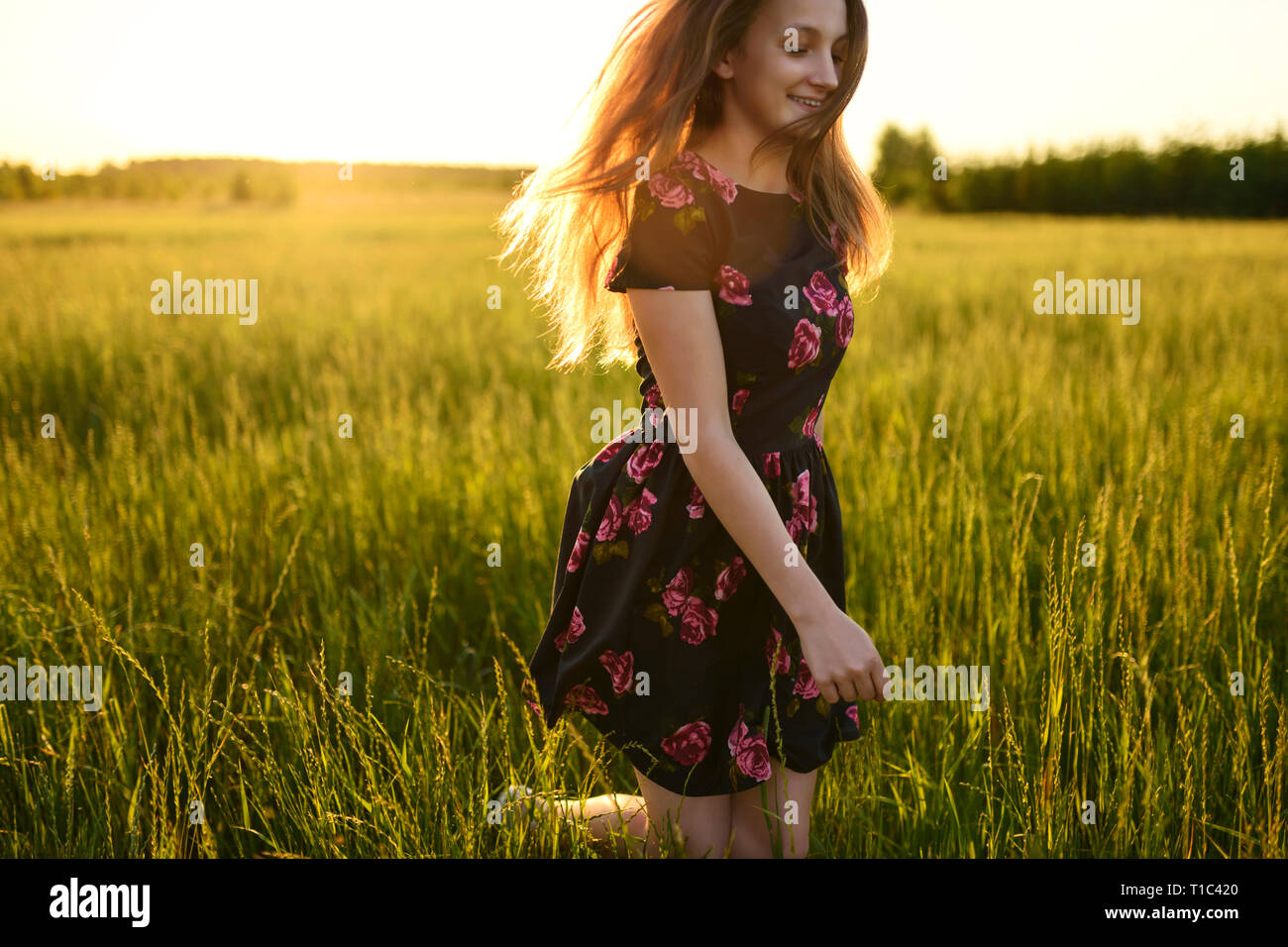 Young girl in flower dress, is running or jumping from happiness on a green meadow. She is smiling and her long hair are flowing on wind, all lit by w Stock Photo