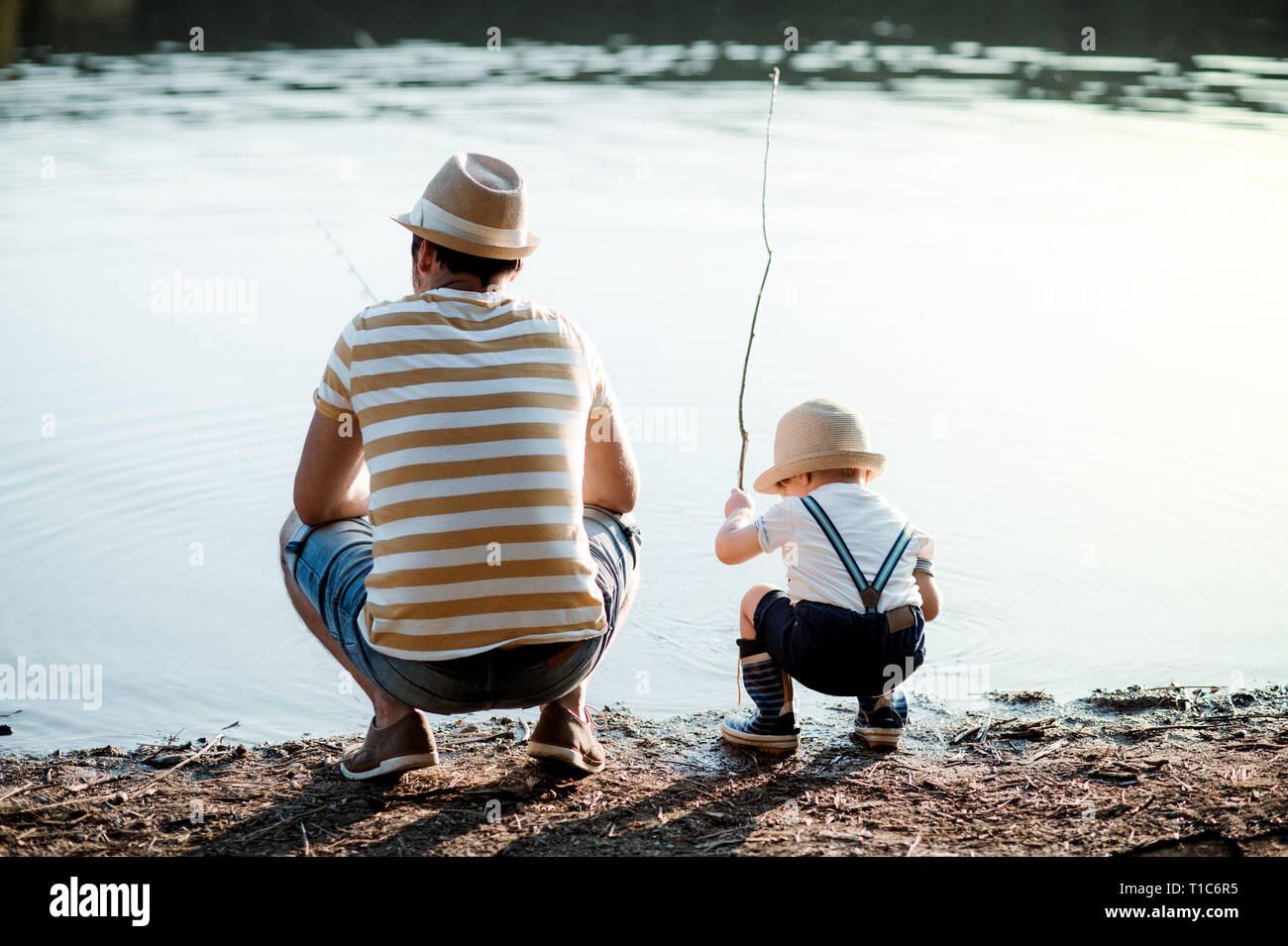 A rear view of mature father with a small toddler son outdoors fishing by a lake. Stock Photo