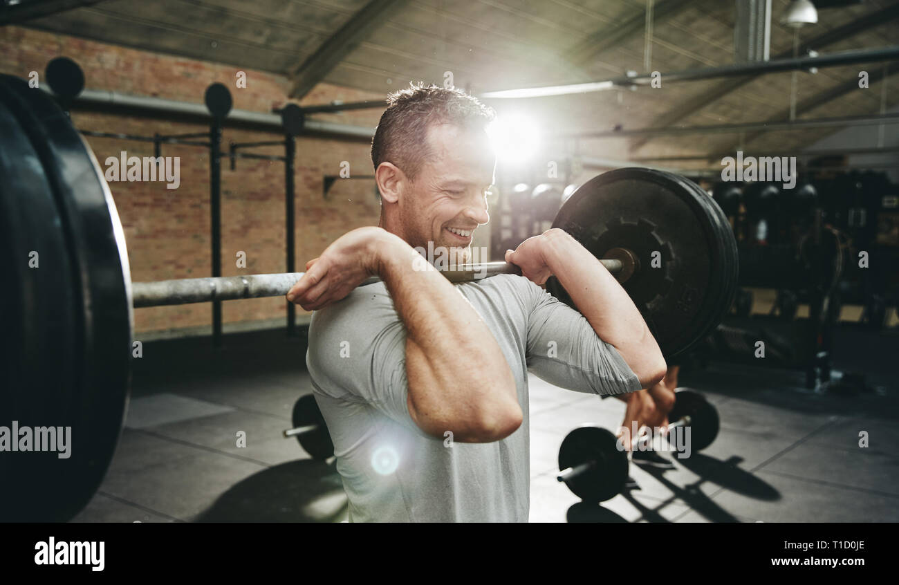 Fit man in sportswear smiling while lifting barbells during a weight training session at the gym Stock Photo