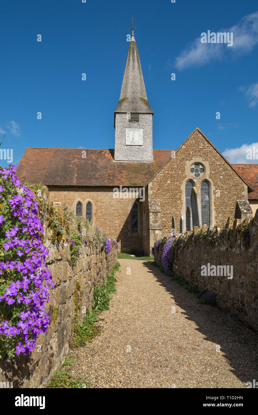 St Michael and All Angels parish church in the village of Thursley, Surrey, UK, on a sunny March day, with spring flowers, purple aubretia or aubrieta Stock Photo