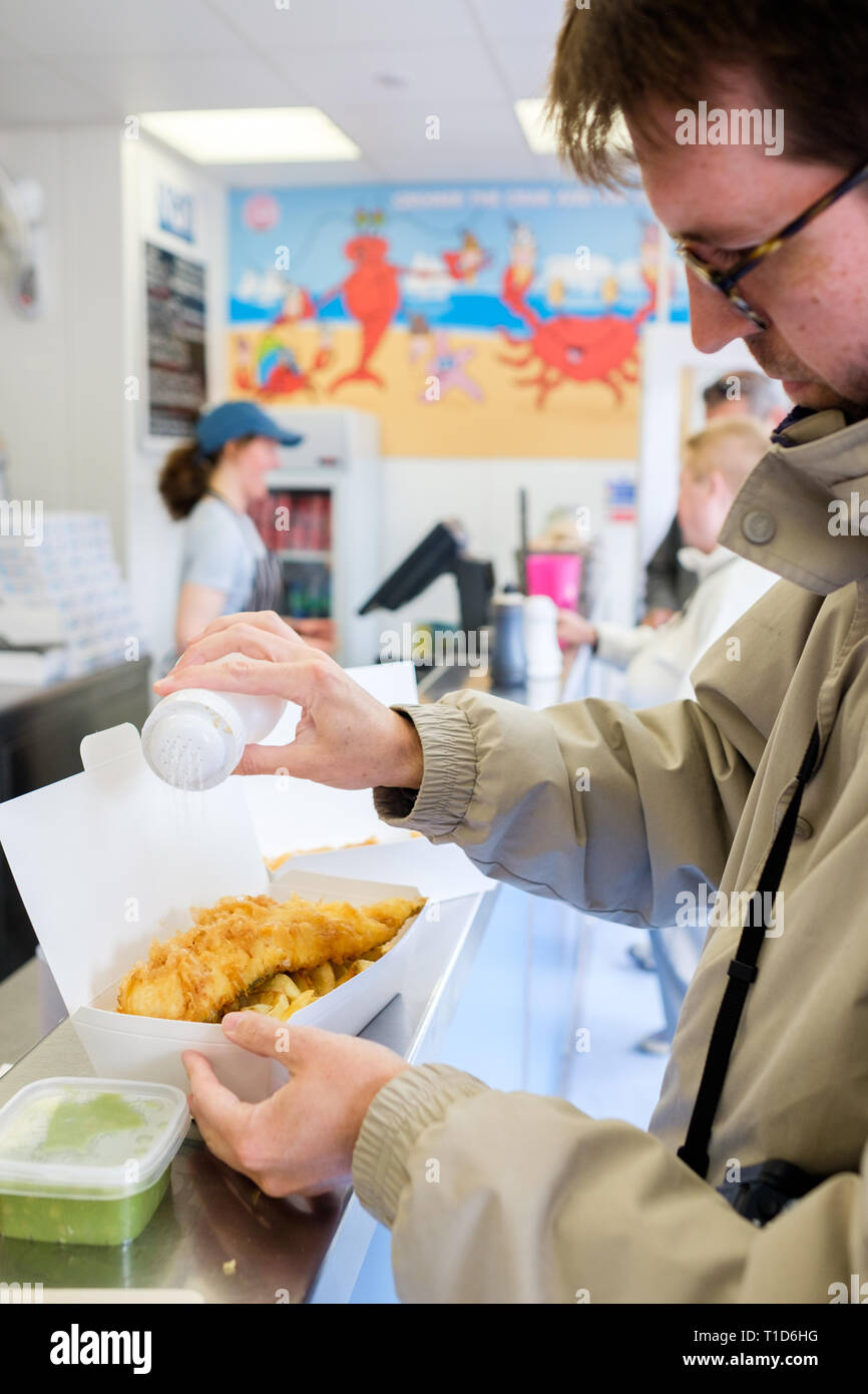 A man puts salt on top of his fish and chips at No1 Fish & Chips Shop in Cromer, North Norfolk, England. Stock Photo