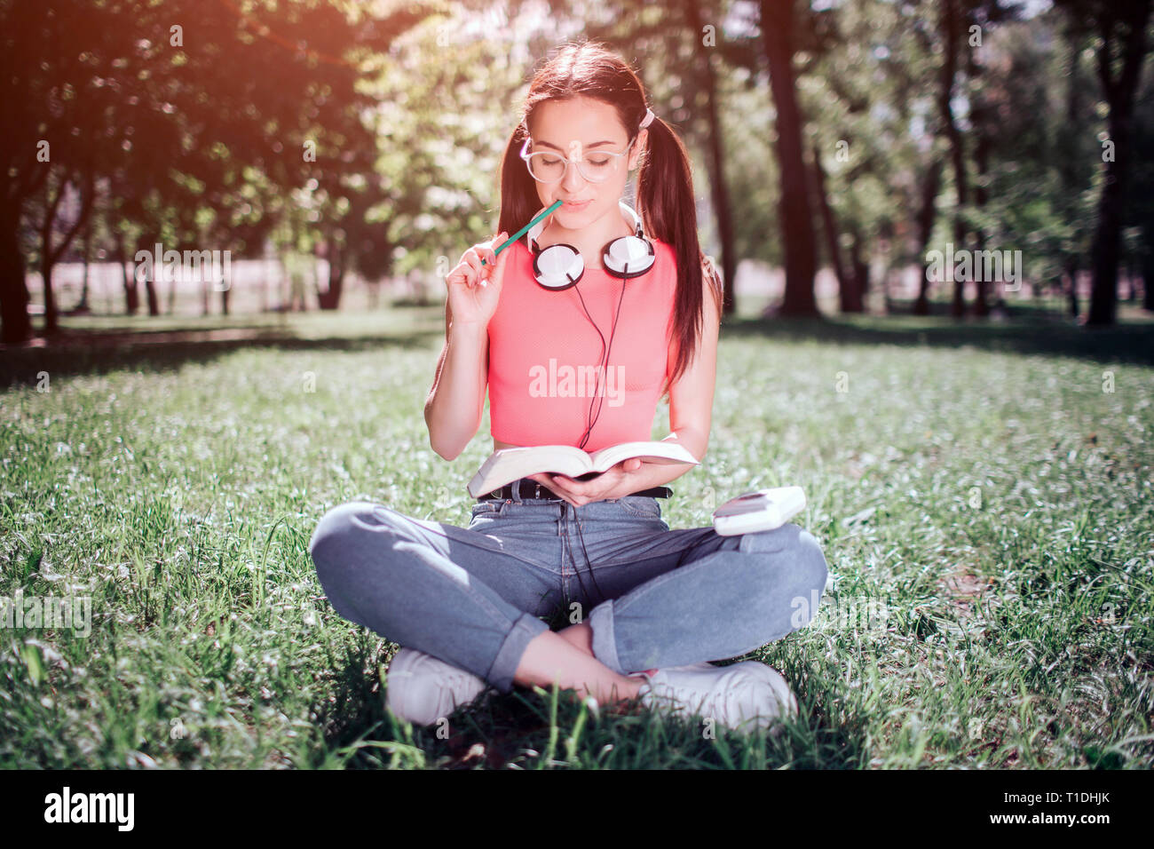 Good and delightful student is sitting on grass and reading a book. She has headphones around her neck. Girl is chewing the end of pencil. Stock Photo