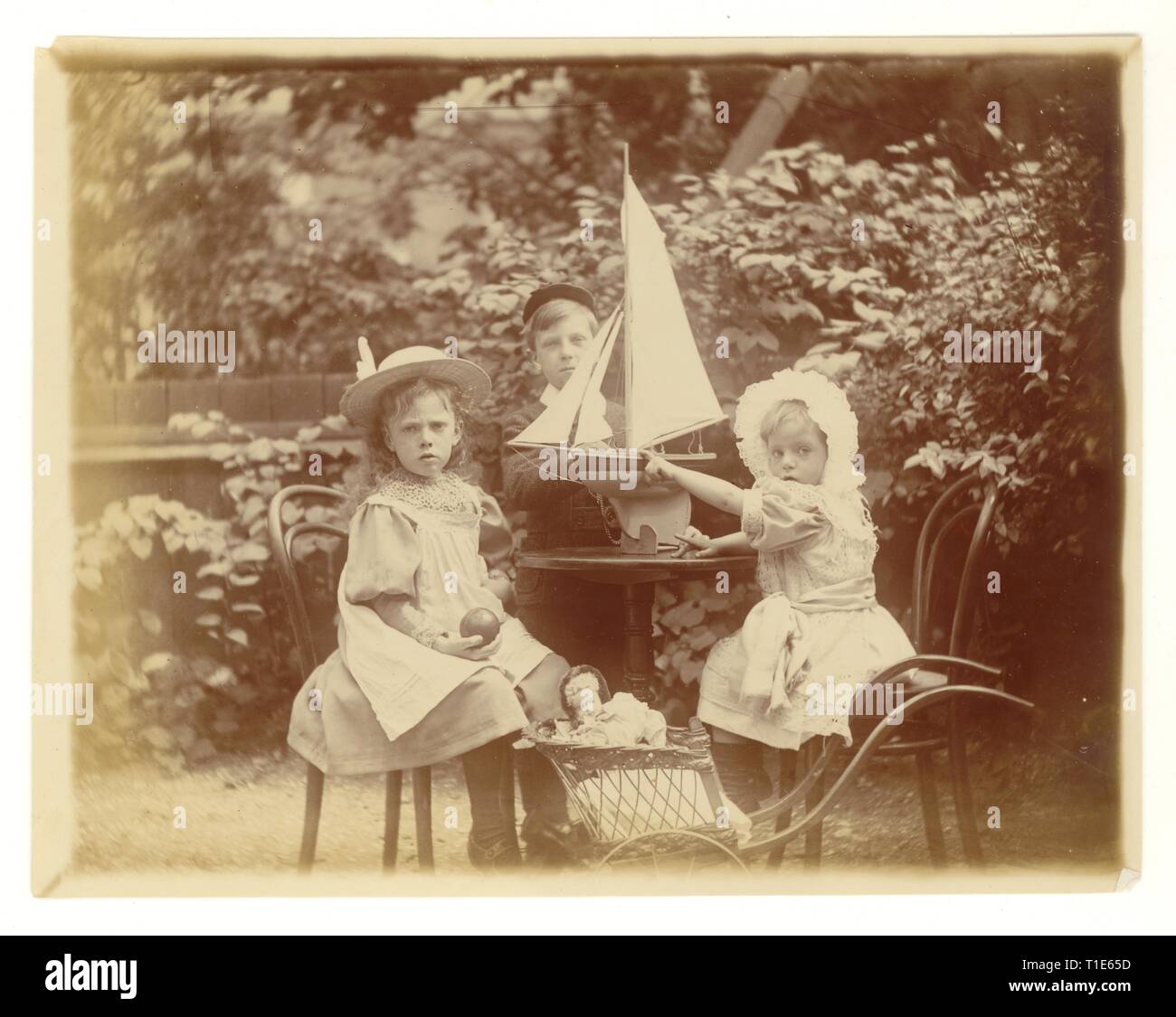 Victorian Photograph of young children in garden wearing hats, (the boy has a fez style hat on) playing with toy yacht and a fashionably dressed doll in a pram, U.K. circa 1894 Stock Photo