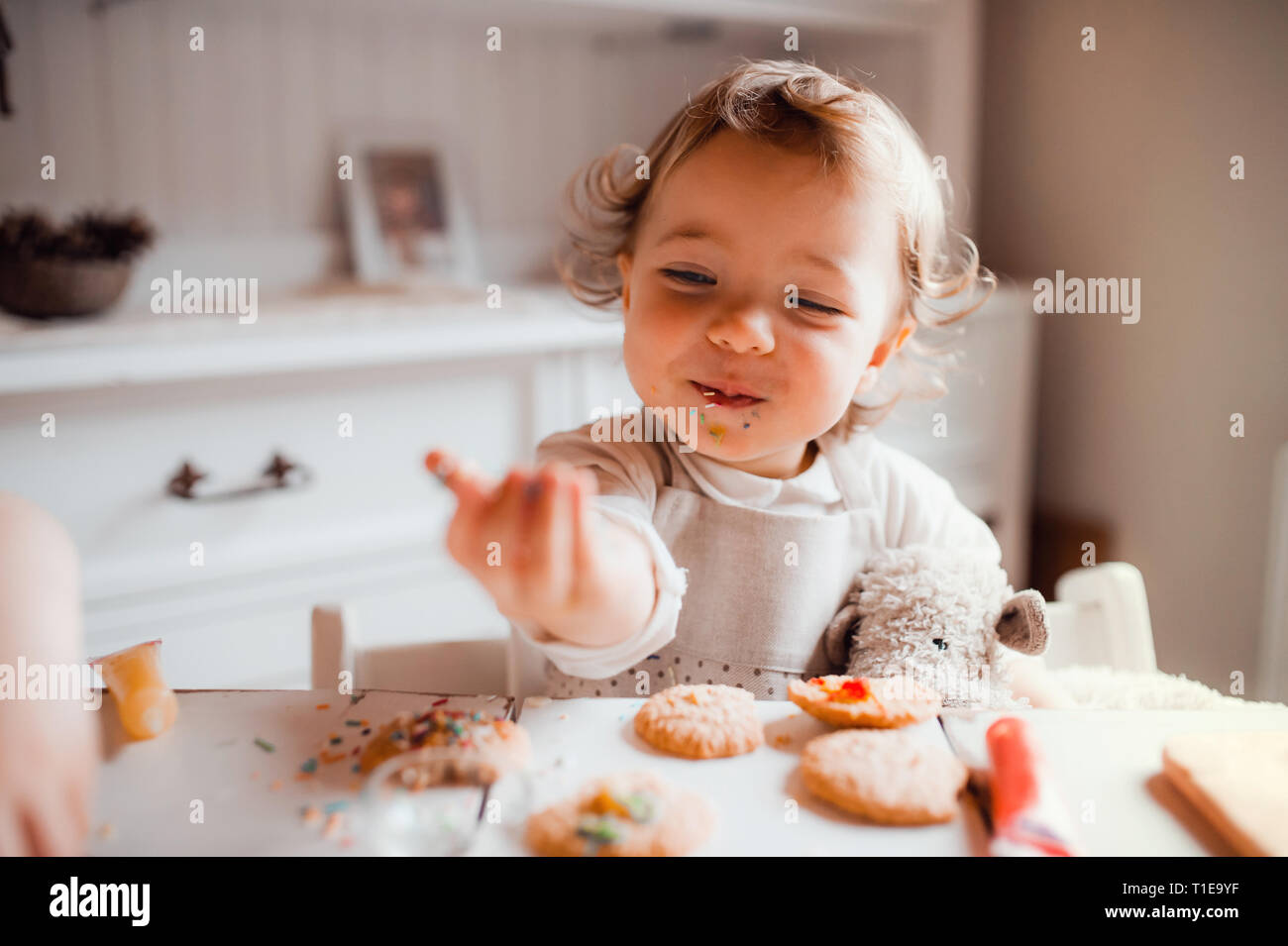 A small toddler girl sitting at the table, decorating cakes at home. Stock Photo