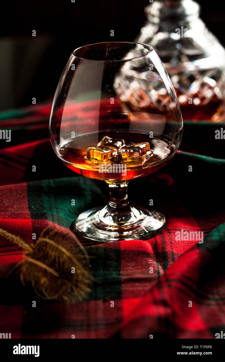 Scottish whiskey in a glass with ice on traditional red with green tartan. Crustal decanter and dry thistle. Selective focus. Stock Photo