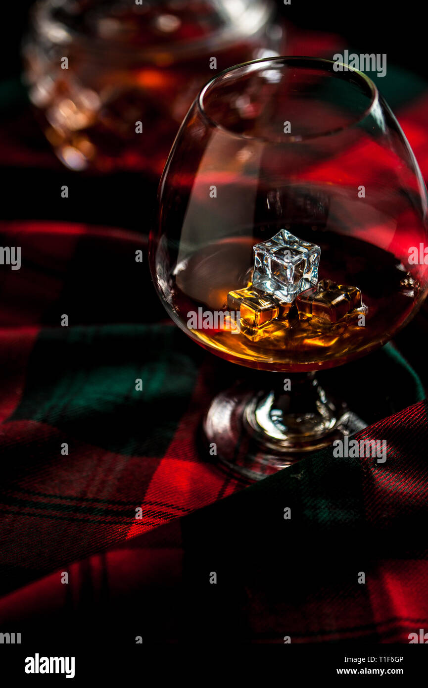 Scottish whiskey in a glass with ice on traditional red with green tartan. Crustal decanter and dry thistle. Selective focus. Stock Photo