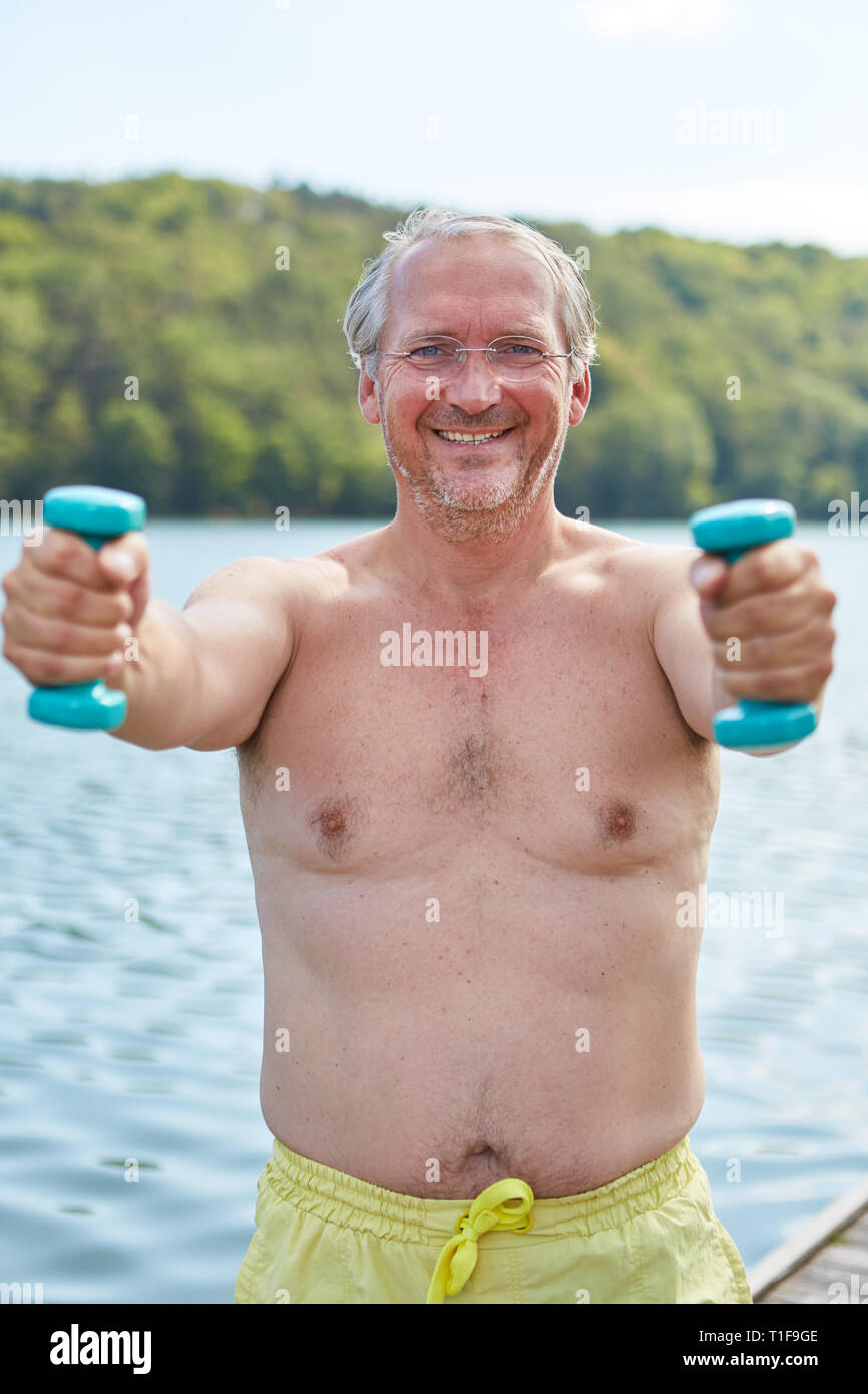 Senior man in swimming trunks on the lake shore is doing weight training for fitness and health Stock Photo