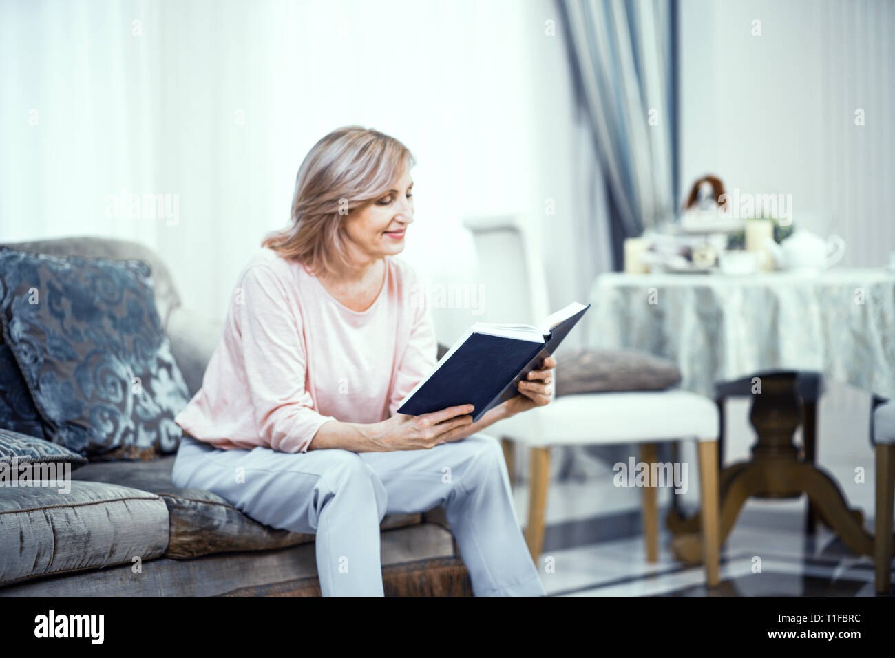 An Elderly Woman in Home Clothes is Sitting on the Sofa With a Book in Her Hands. Stock Photo