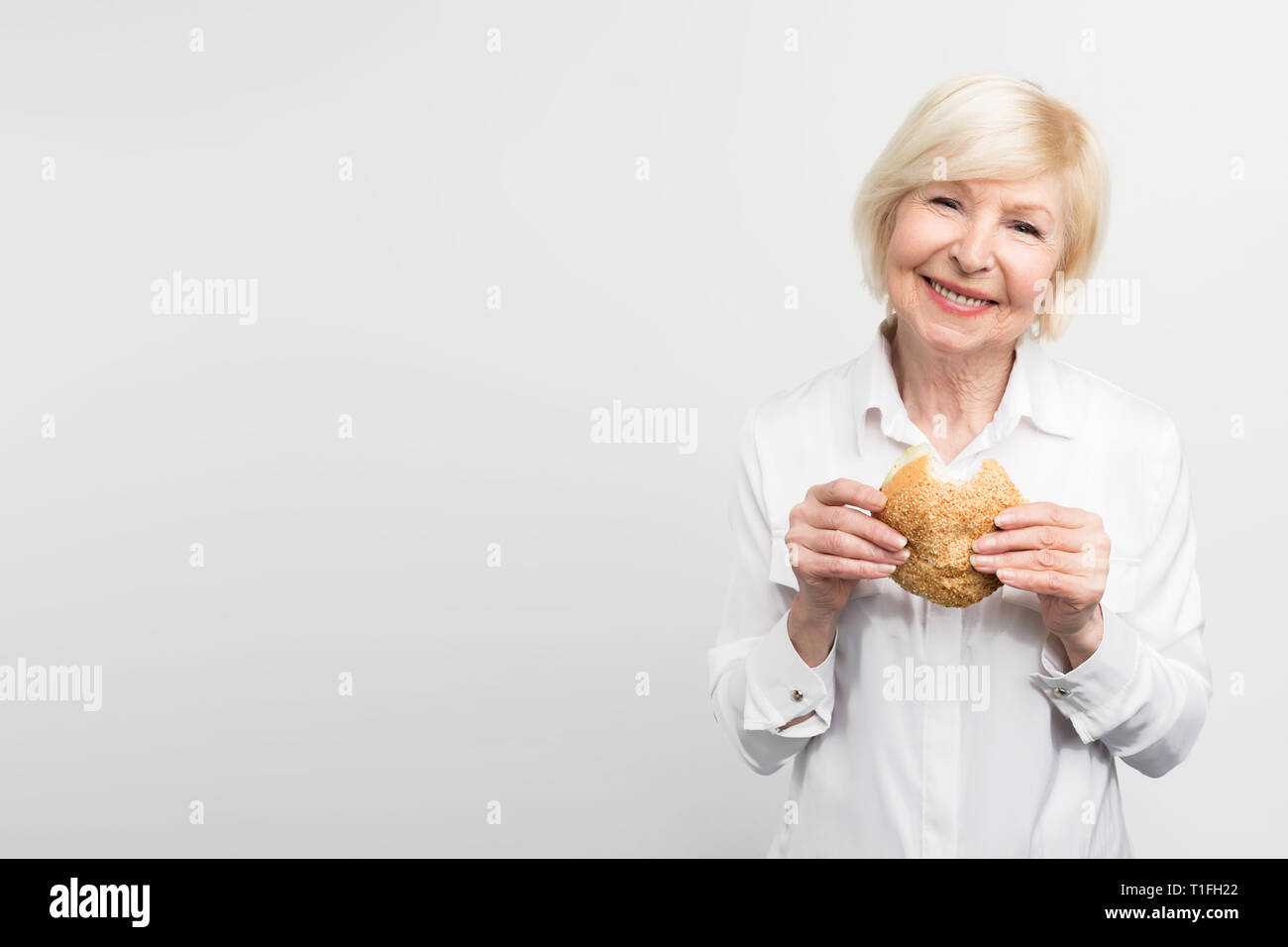 Old but satisfied woman is holding a burger in her hands. She has just made one bite. This lady likes the taste of this meal. Sometimes she likes to Stock Photo