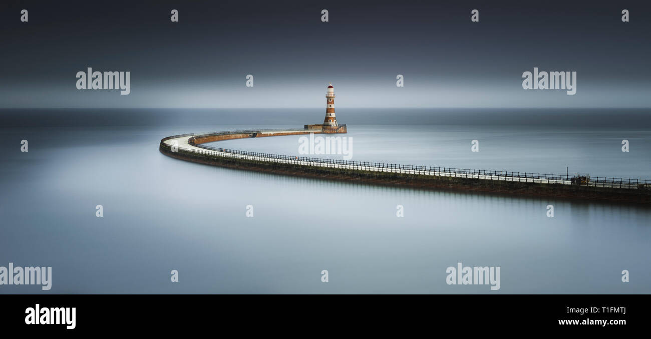 A moody Fine Art colour long exposure of Roker Pier at sunset with the long sweeping path giving a soft mirror reflection in the North Sea Stock Photo