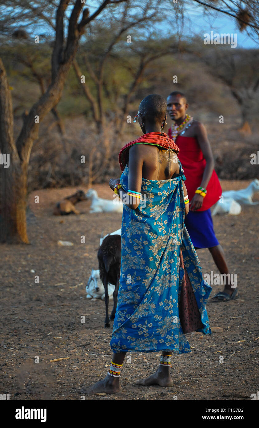 Sunset glow on a typical village scene, Samburu, Kenya, Africa.  Woman in traditional dress with back to camera looks towards male villager. Stock Photo