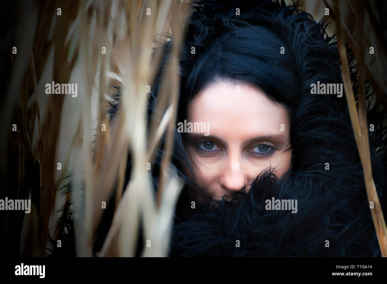 Close-up outdoor portrait of a beautiful woman with black hair looking through tall straw like blades of leaves Stock Photo