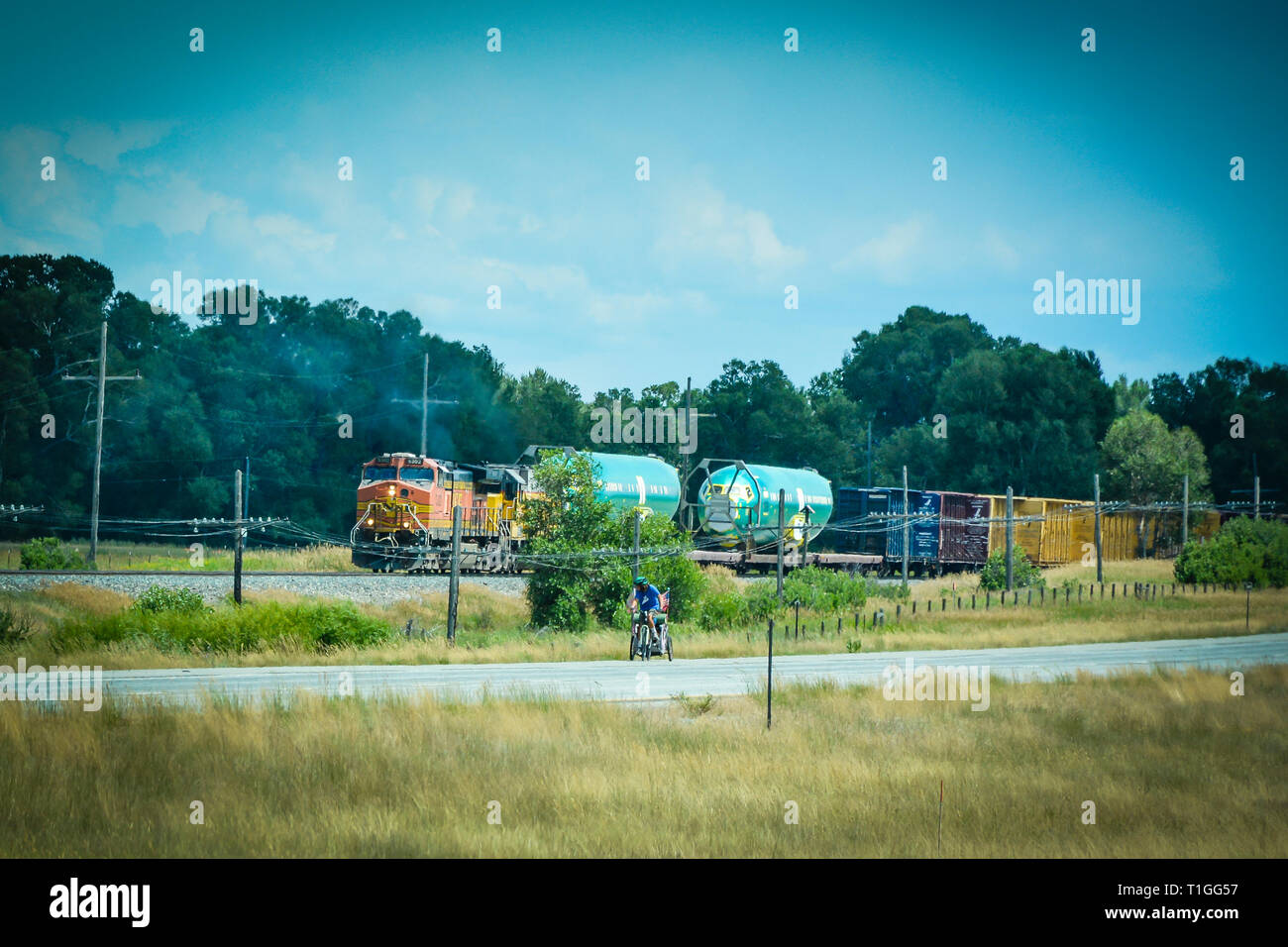A cyclist on a road has a look at BNSF train that is about to catch up and pass him on the nearby track, in a rural area of Southwestern Montana, USA Stock Photo