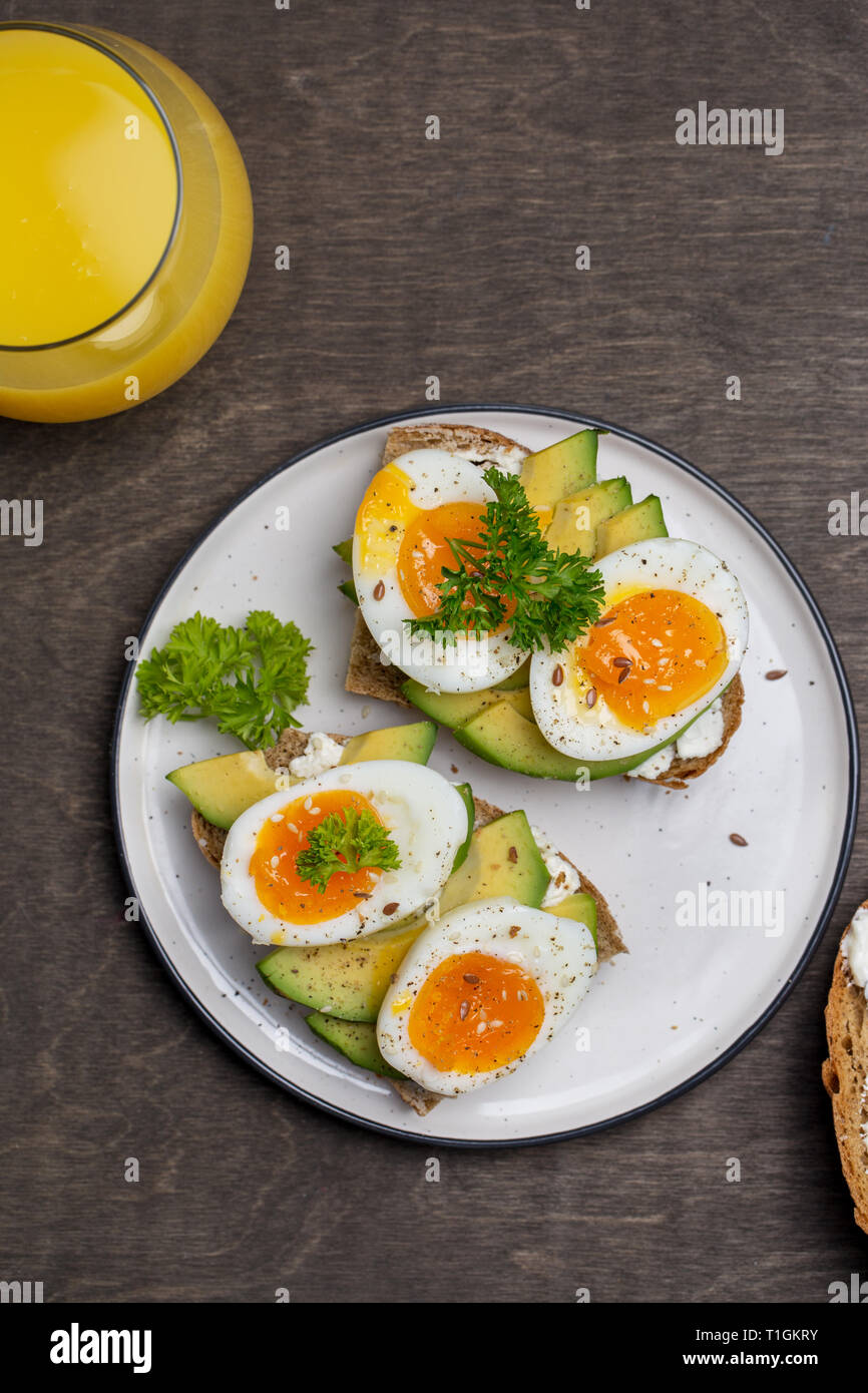 Two tasty sandwiches with avocado and boiled egg on white plate, grain bread with cream cheese and glass of orange juice near on wooden background Stock Photo