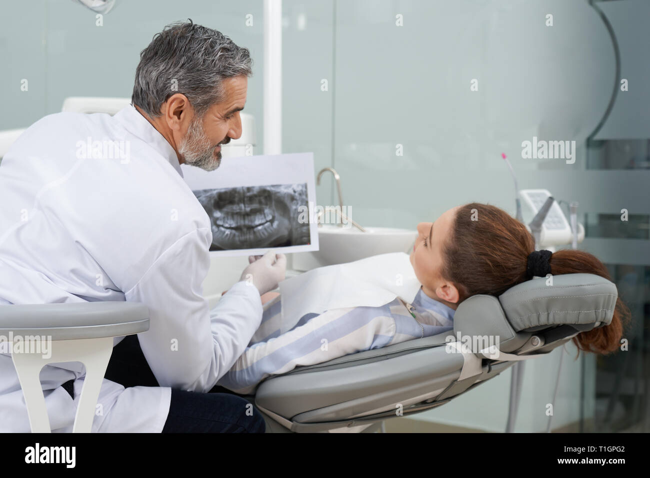 Dentist showing to client dental xray of teeth and jaw. Mature, bearded man wearing white