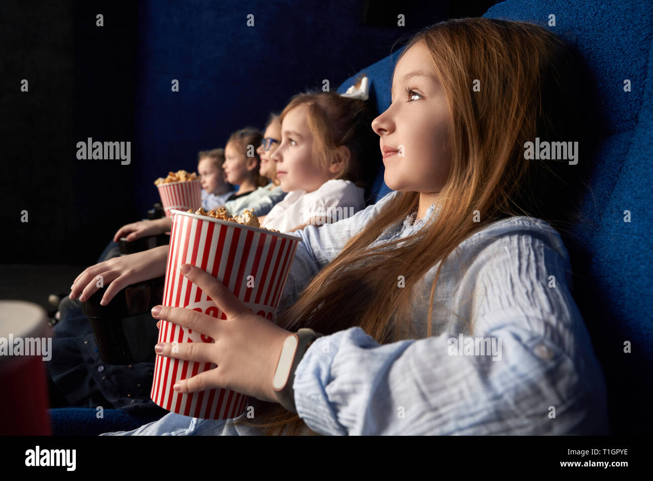 Pretty, little girl holding popcorn bucket, sitting with friends in cinema, in comfortable chairs. Children watching cartoon or movie. Leisure, entertainment. Stock Photo