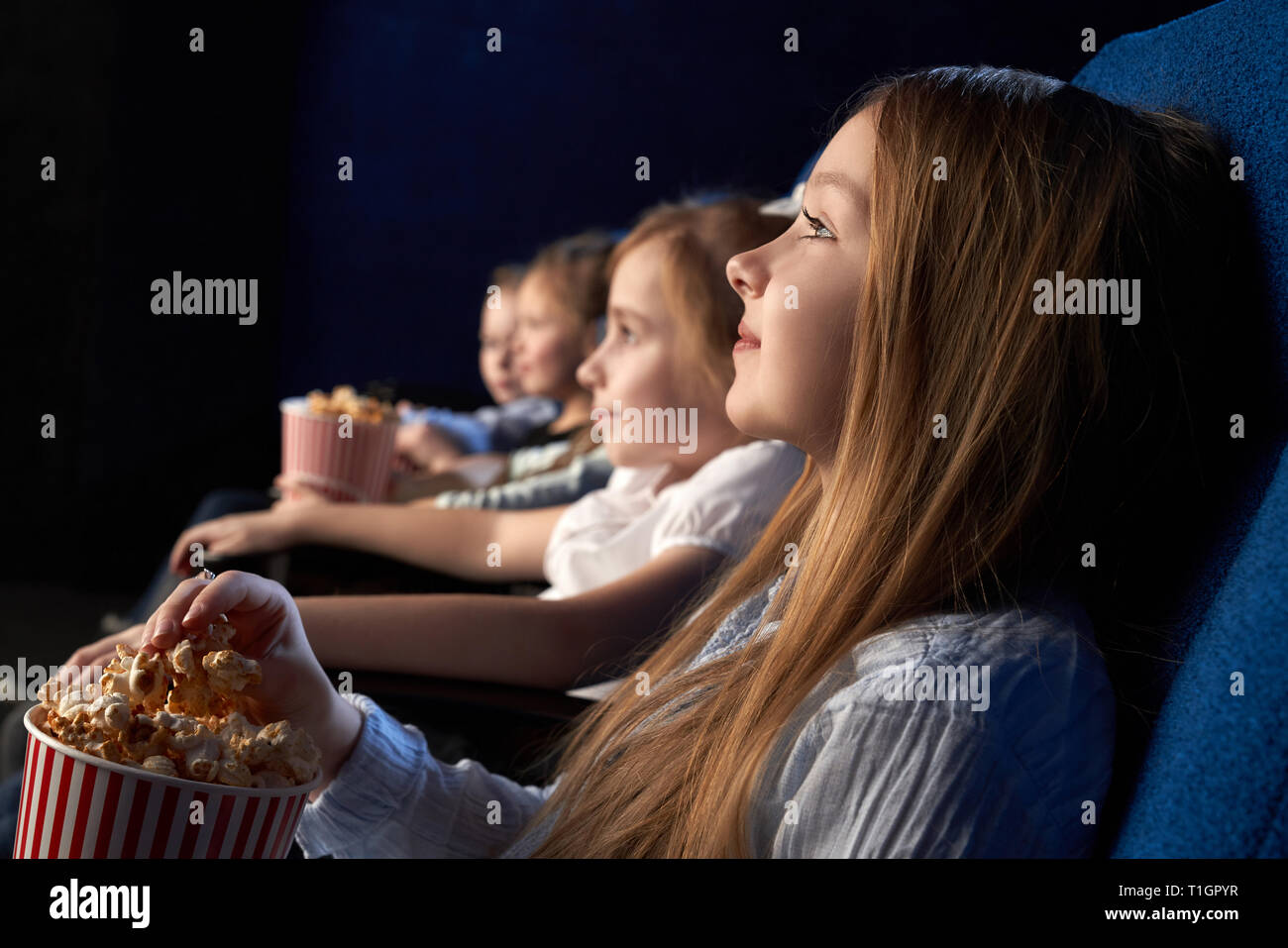 Kids sitting in comfortable chairs of movie theatre, watching attentively movie or cartoon. Beautiful girl holding popcorn bucket, enjoying film premiere. Stock Photo