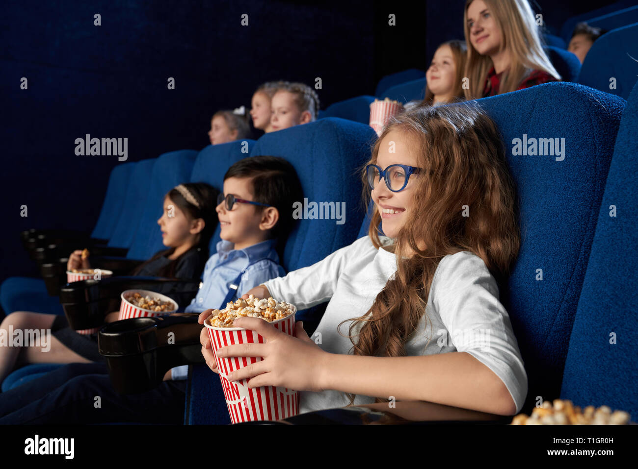 Happy, joyful kids sitting in dark blue, comfortable chairs in modern cinema theatre, watching movie or cartoon. Children enjoying film, smiling, holding buckets with popcorn. Stock Photo