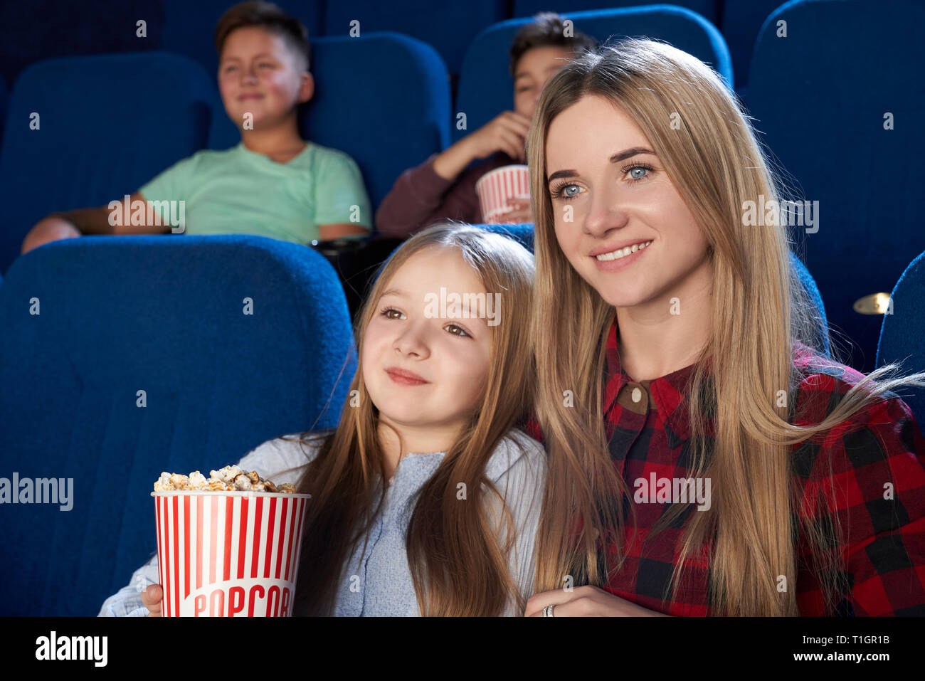 Beautiful woman sitting with little girl in cinema, enjoying premiere of film. Cheerful mother and daughter watching movie, girl holding popcorn bucket. Stock Photo