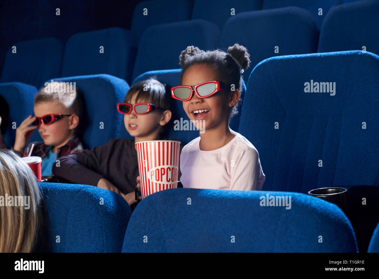 Cheerful, joyful children sitting in cinema theatre, enjoying movie or cartoon. Kids wearing in 3D glasses. Cute little girl smiling, holding popcorn bucket. Stock Photo