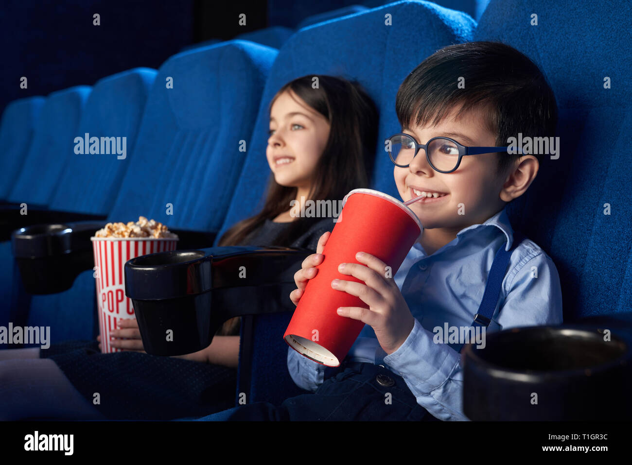 Two joyful, happy kids sitting in cinema theatre, watching cartoon or movie. Cheerful, handsome boy holding red paper cup, sipping fizzy drink, pretty girl eating popcorn. Stock Photo