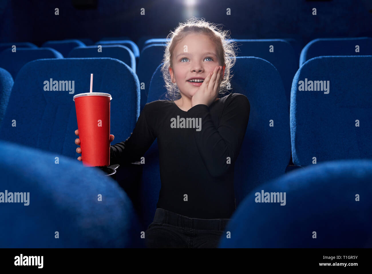 Little, cute girl watching attentively movie in cinema theatre, holding red paper cup with fizzy drink in hand. Fascinated kid holding hand on cheek. Stock Photo