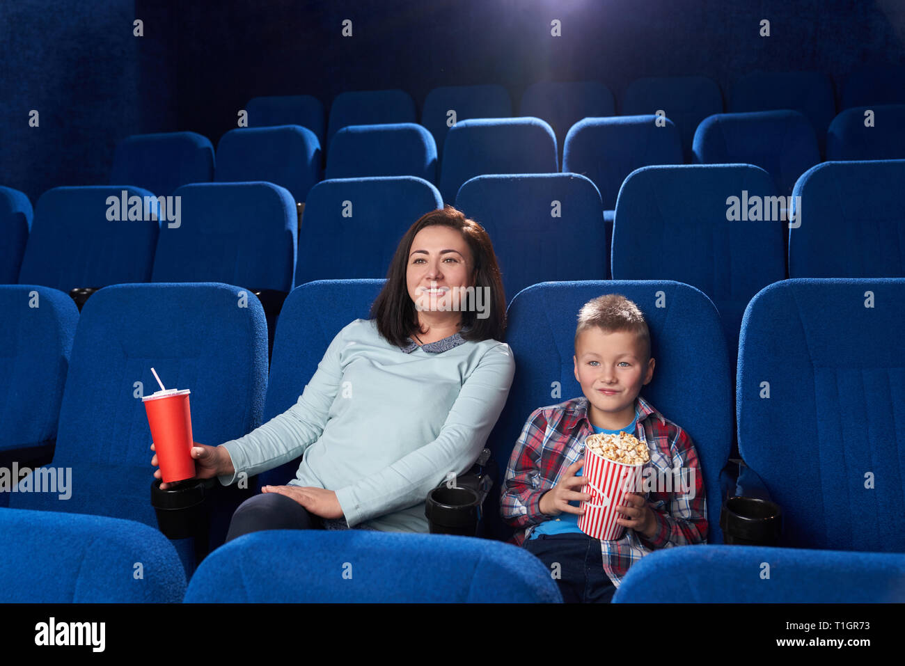 Mother and son sitting together in cinema theatre. Happy, cheerful boy and beautiful woman holding popcorn bucket, red paper cup with fizzy drink. Family time, entertainment. Stock Photo