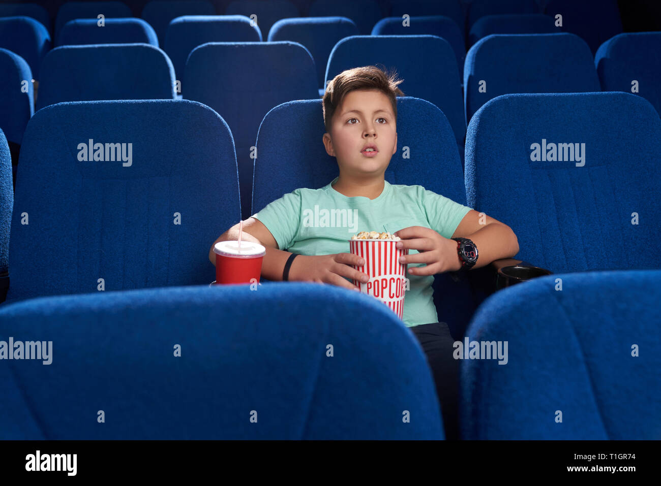 Teenager sitting in movie theatre in comfortable dark blue chair. Handsome boy holding popcorn bucket, red paper cup with fizzy drink. Concept of entertainment and leisure. Stock Photo