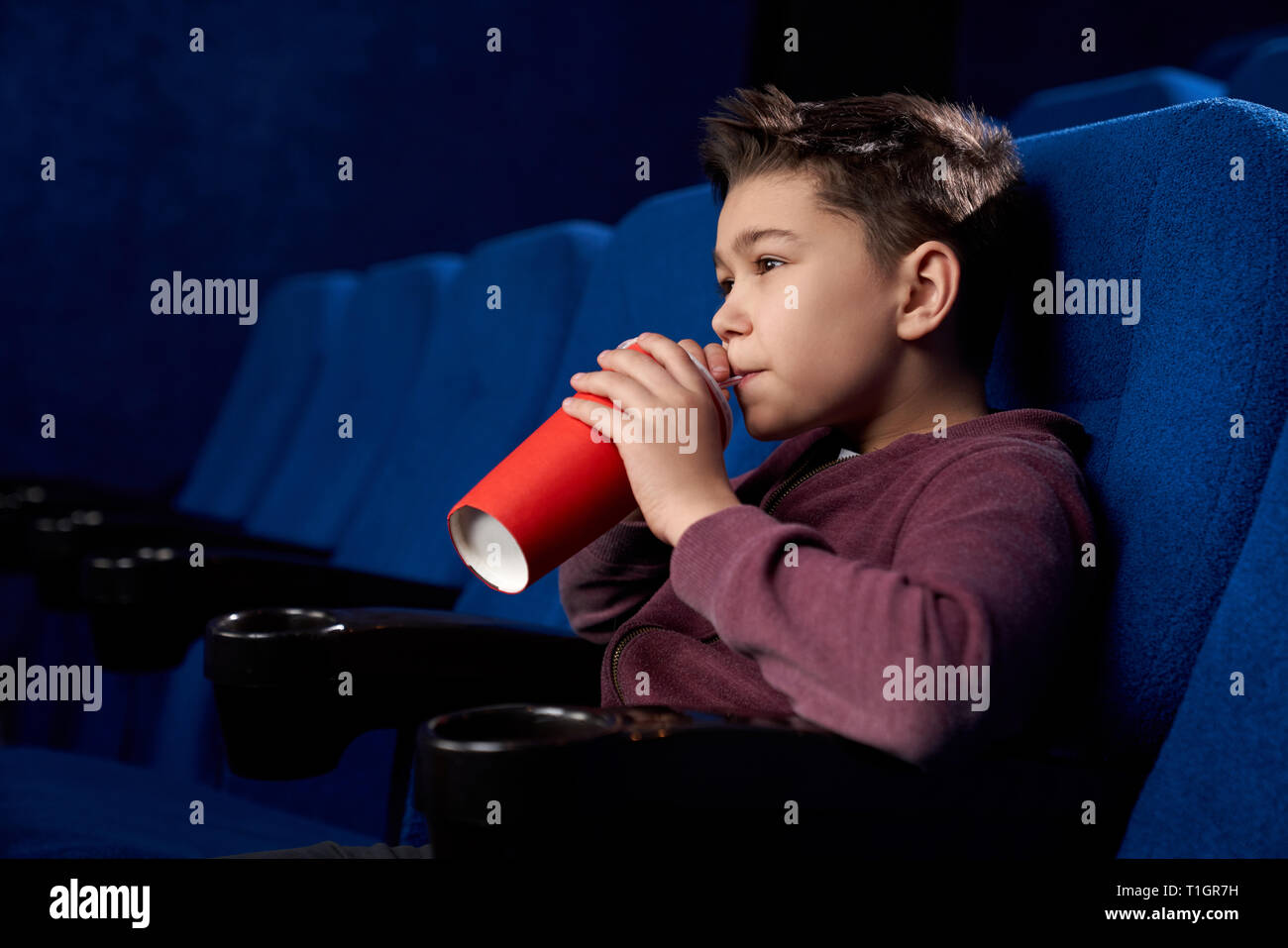 Side view of boy watching film in cinema theatre, sipping, drinking fizzy drink with straw. Teenager holding red paper cup, enjoying movie. Stock Photo