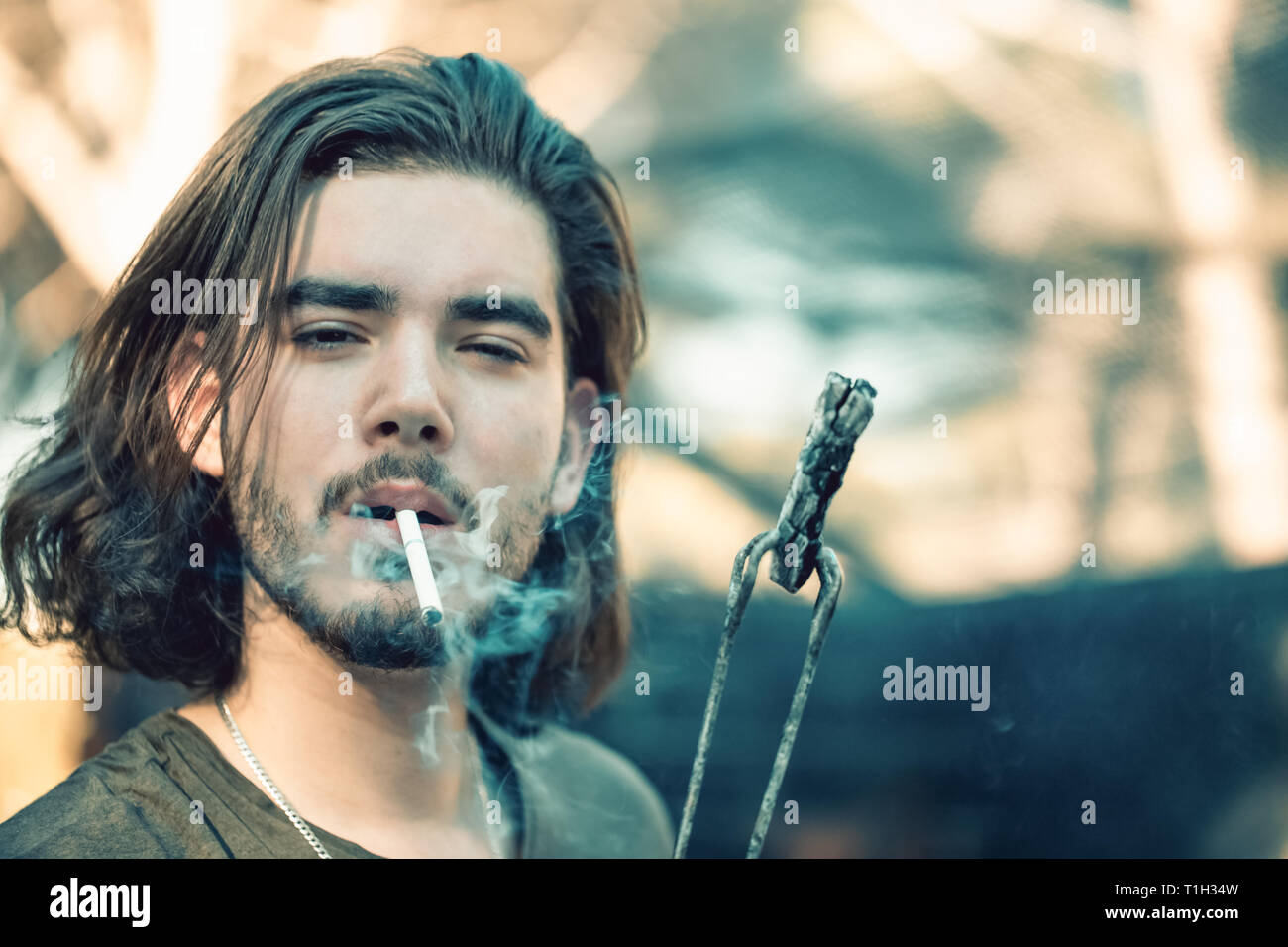 Portrait of a handsome young man with a beard, lighting a cigarette from charcoal, holding his by barbecue tongs. Stock Photo