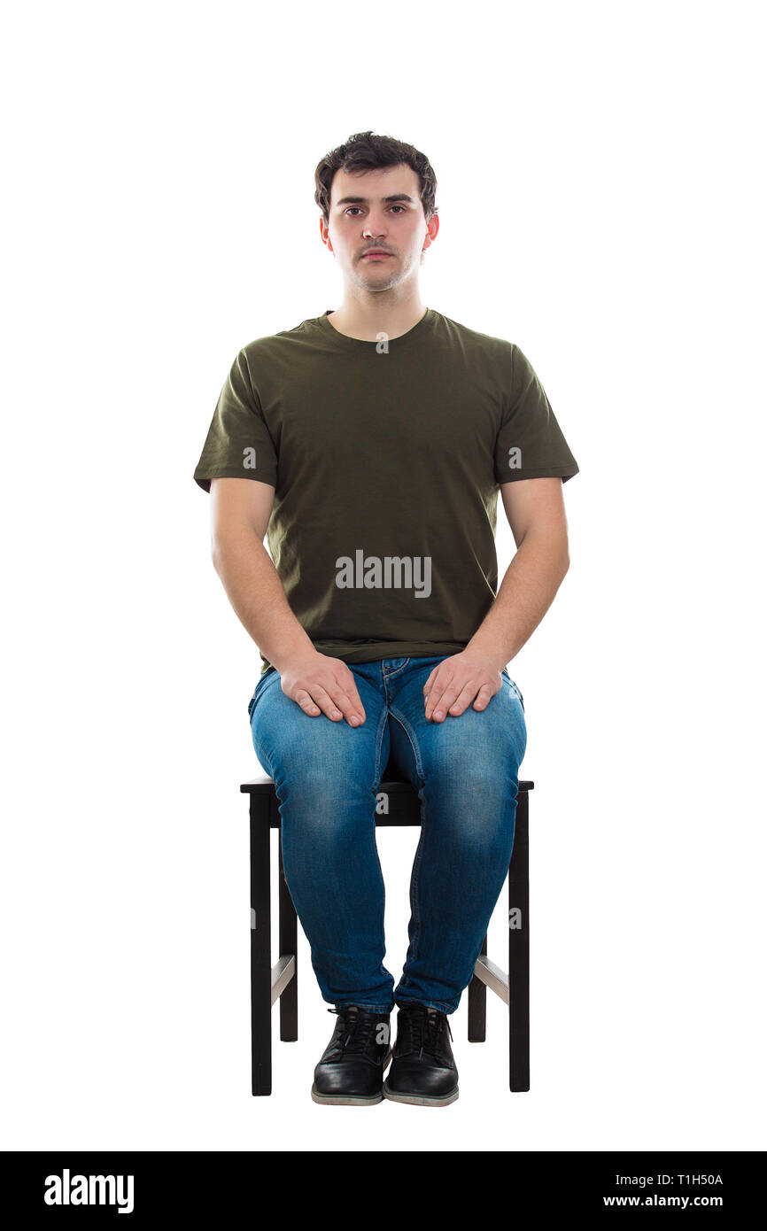 Full length portrait of casual young man sitting on a chair, hands on knees, looking serious to camera isolated over white background. Stock Photo